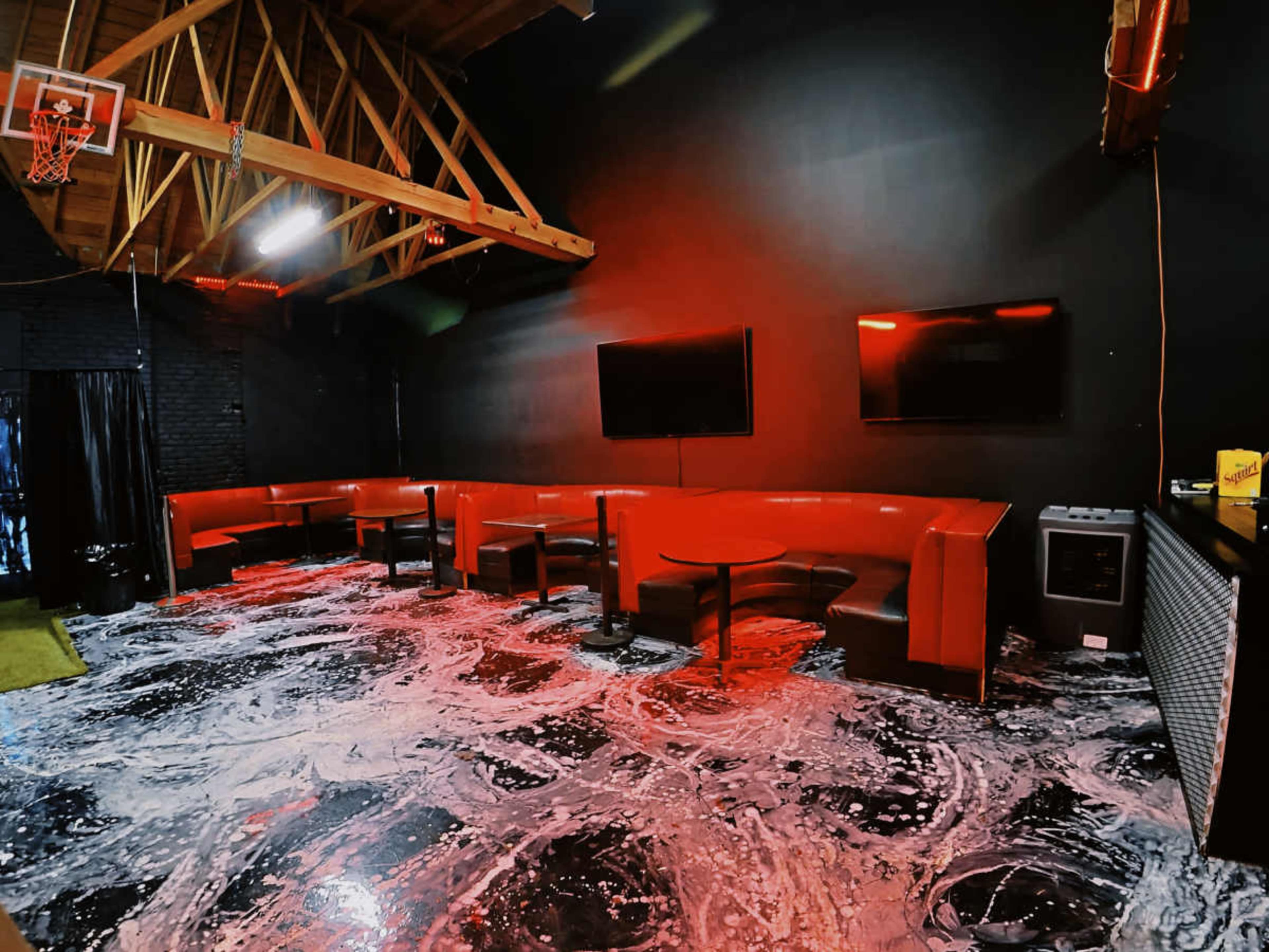 The scene features a dimly lit lounge area with red seating, two wall-mounted televisions, and a glossy floor design resembling swirling patterns.