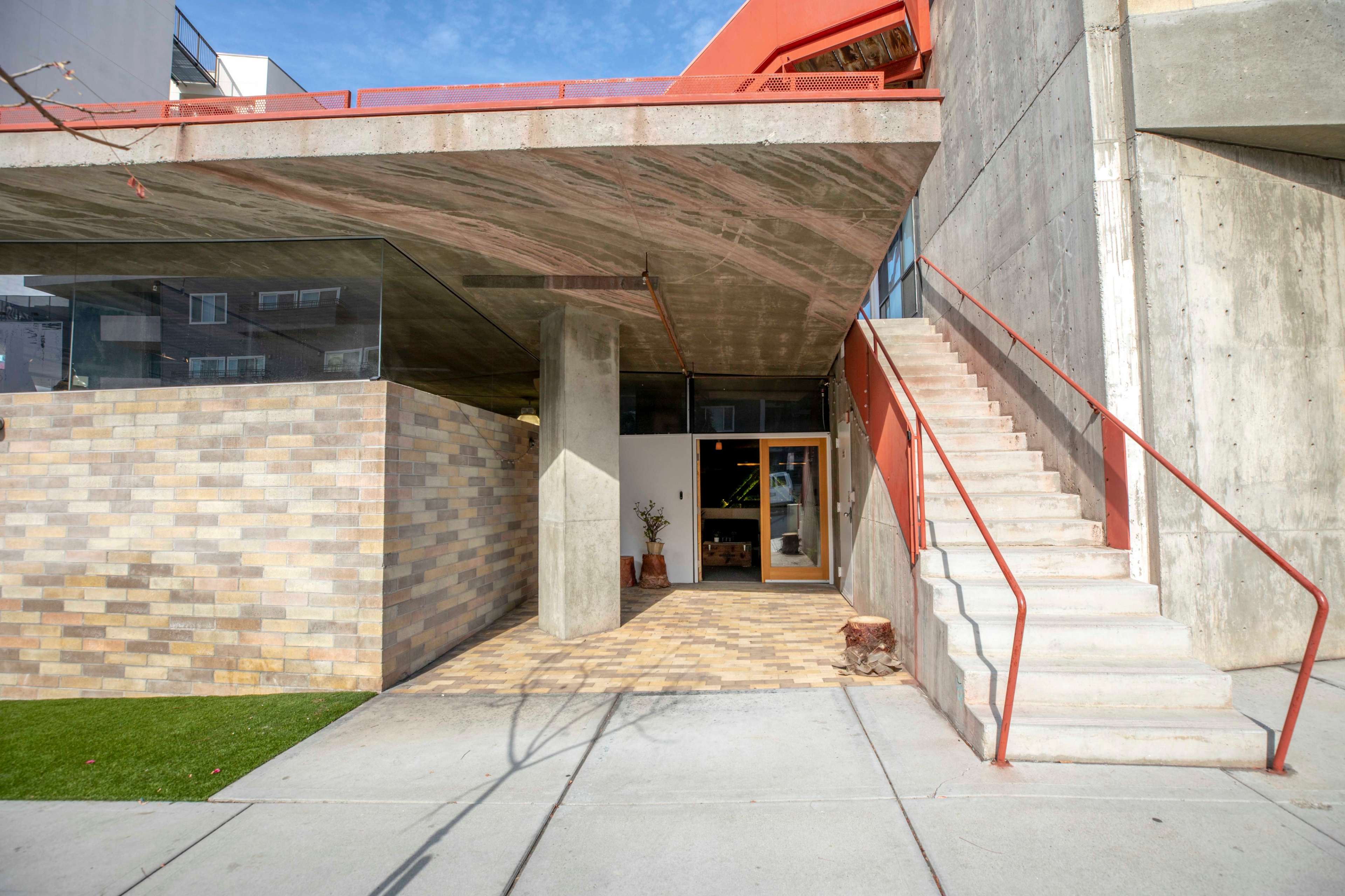 The image shows a modern building entrance with a concrete facade, glass walls, and a staircase leading to an upper level.