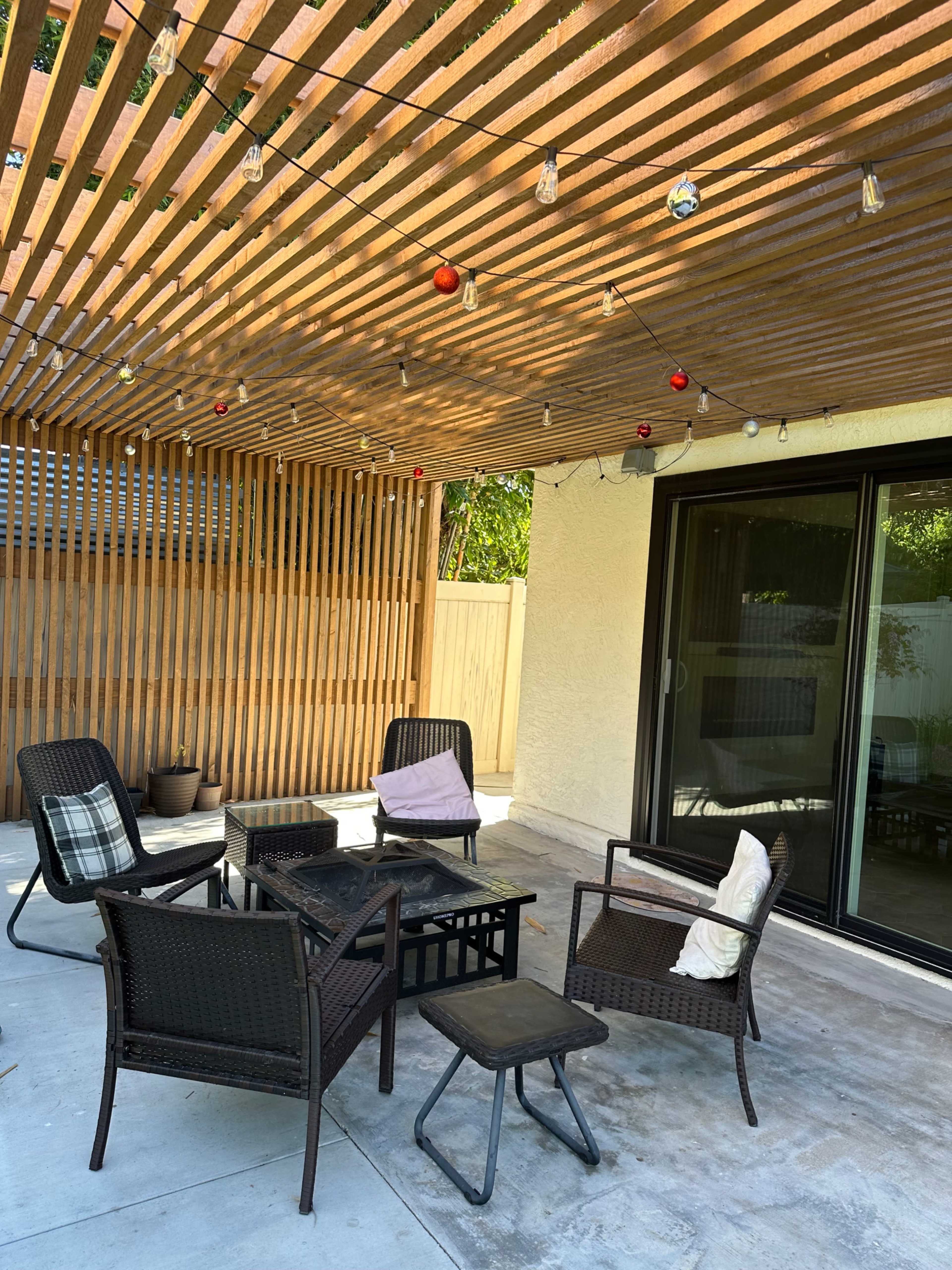 A backyard patio area with four chairs around a black coffee table, beneath a wooden slatted roof decorated with string lights and small ornaments.