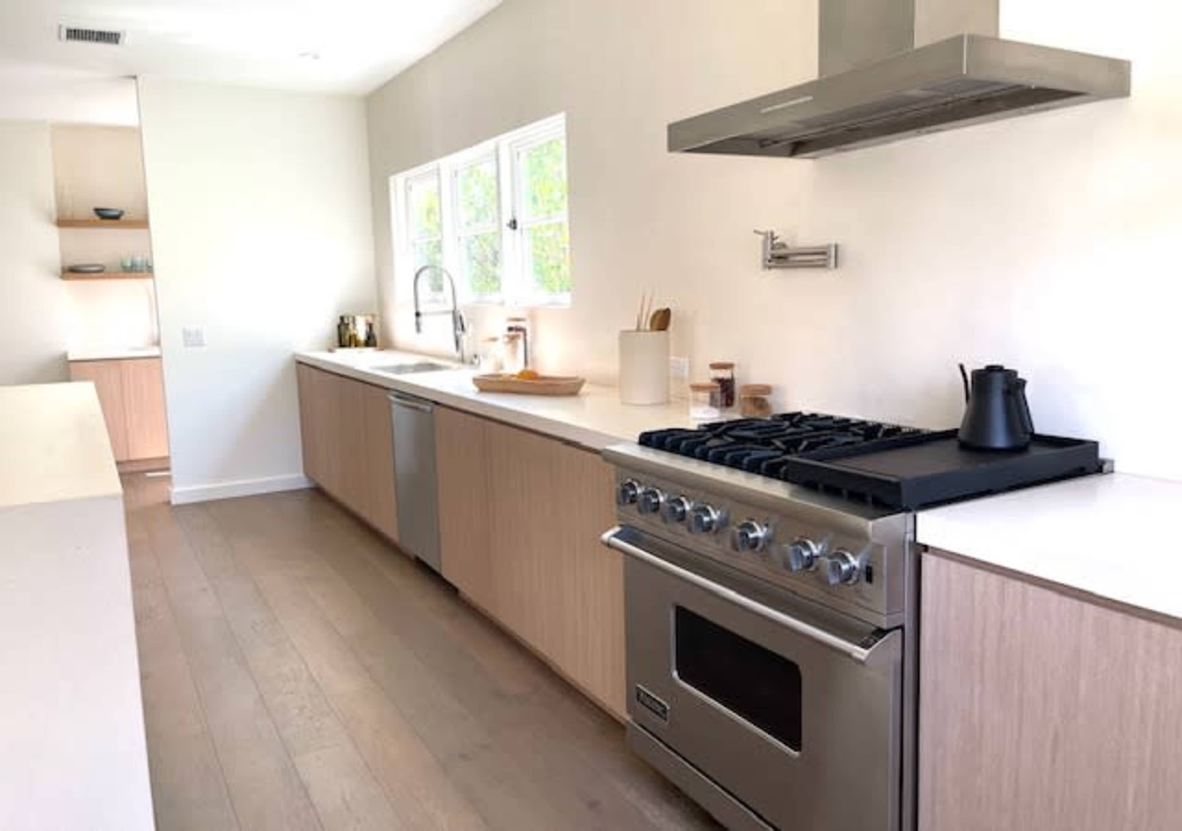 The image shows a modern kitchen featuring light wood cabinetry, a stainless steel gas range, and a large window above the sink.