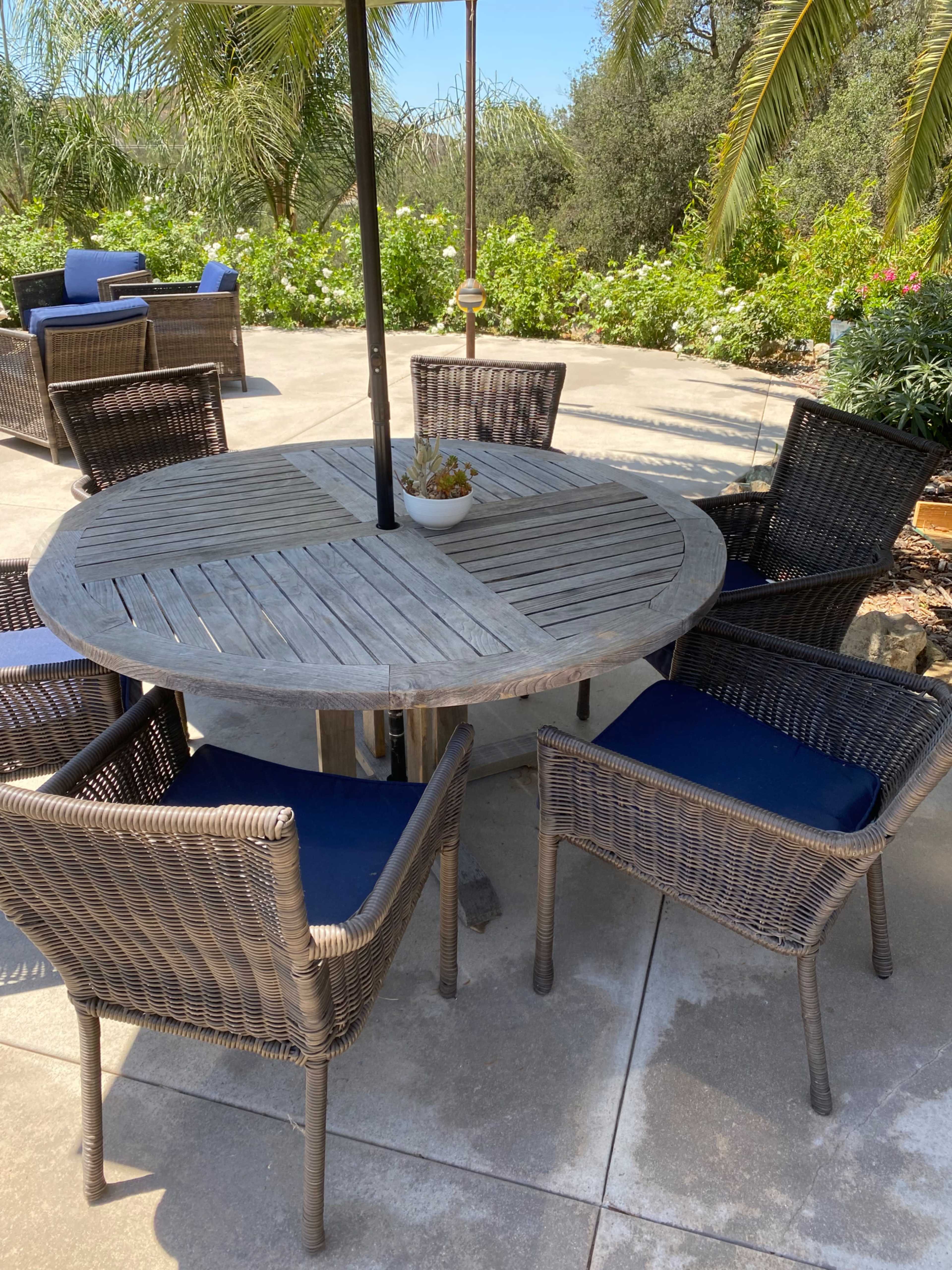 A round wooden table with an umbrella is surrounded by four wicker chairs on a patio beneath palm trees and lush greenery.