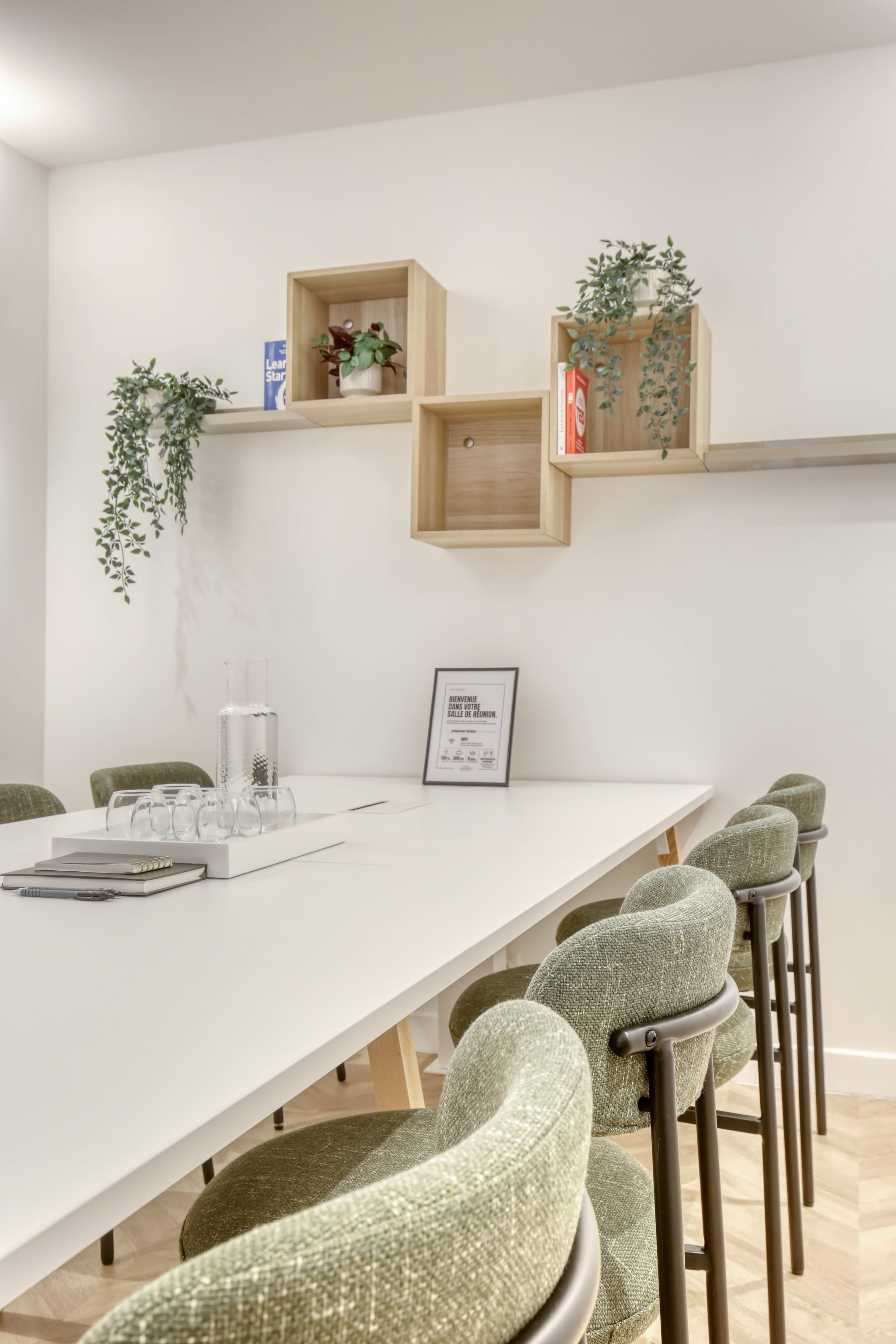 A long white table with green upholstered chairs is set in a modern meeting room decorated with wooden shelves and plants on the walls.