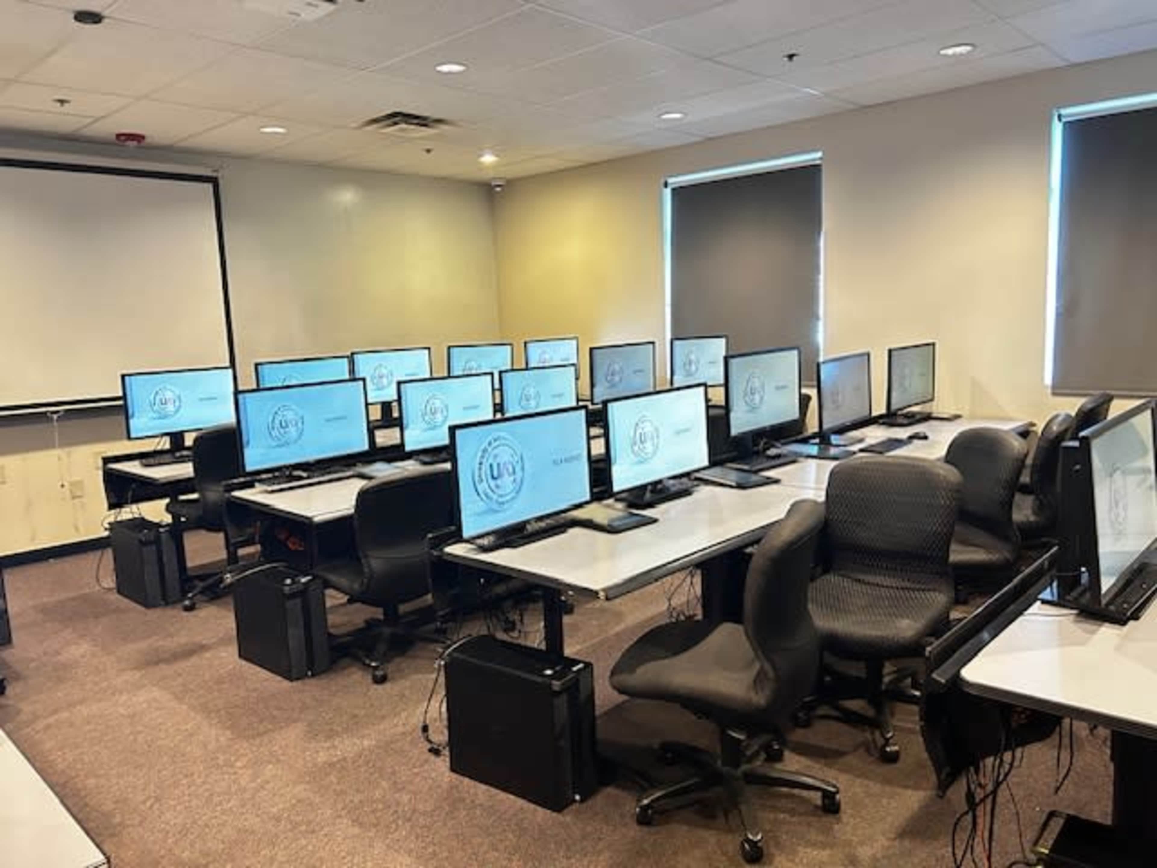 A classroom is arranged with multiple computer stations, each featuring a monitor and chair, facing a presentation screen.