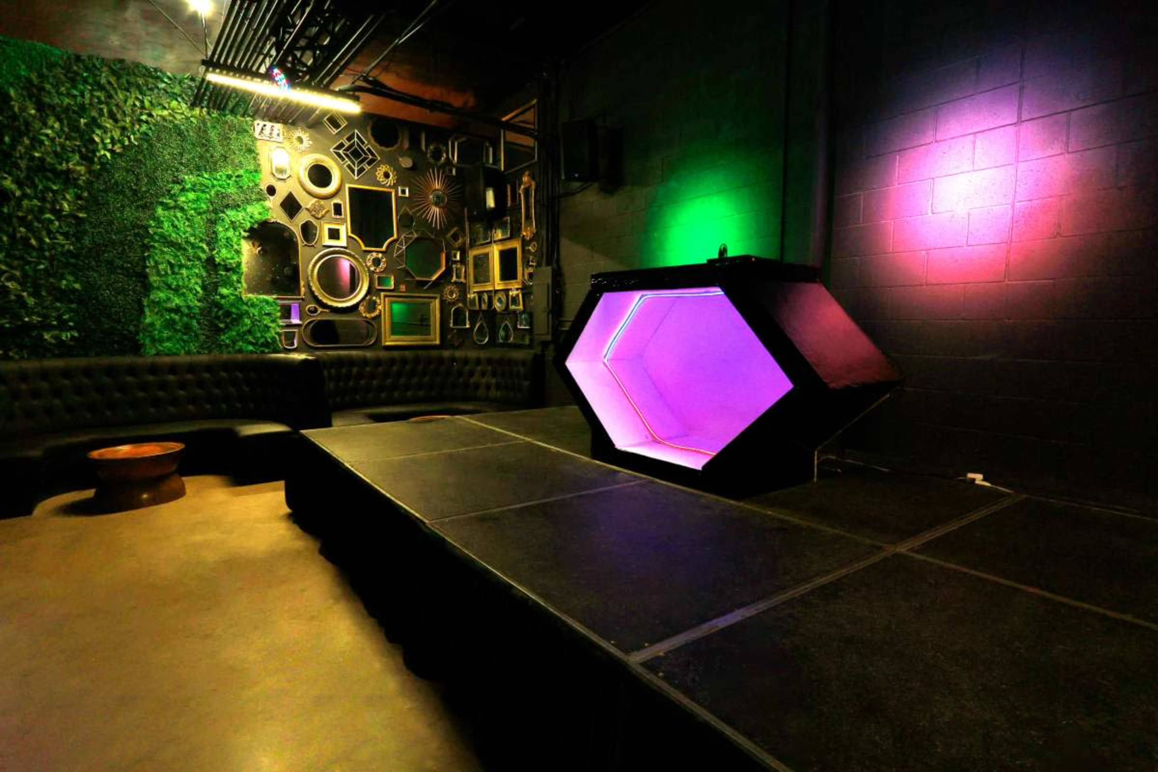 The image shows a small stage in a venue with a geometric light display and a wall decorated with various framed artworks and greenery.