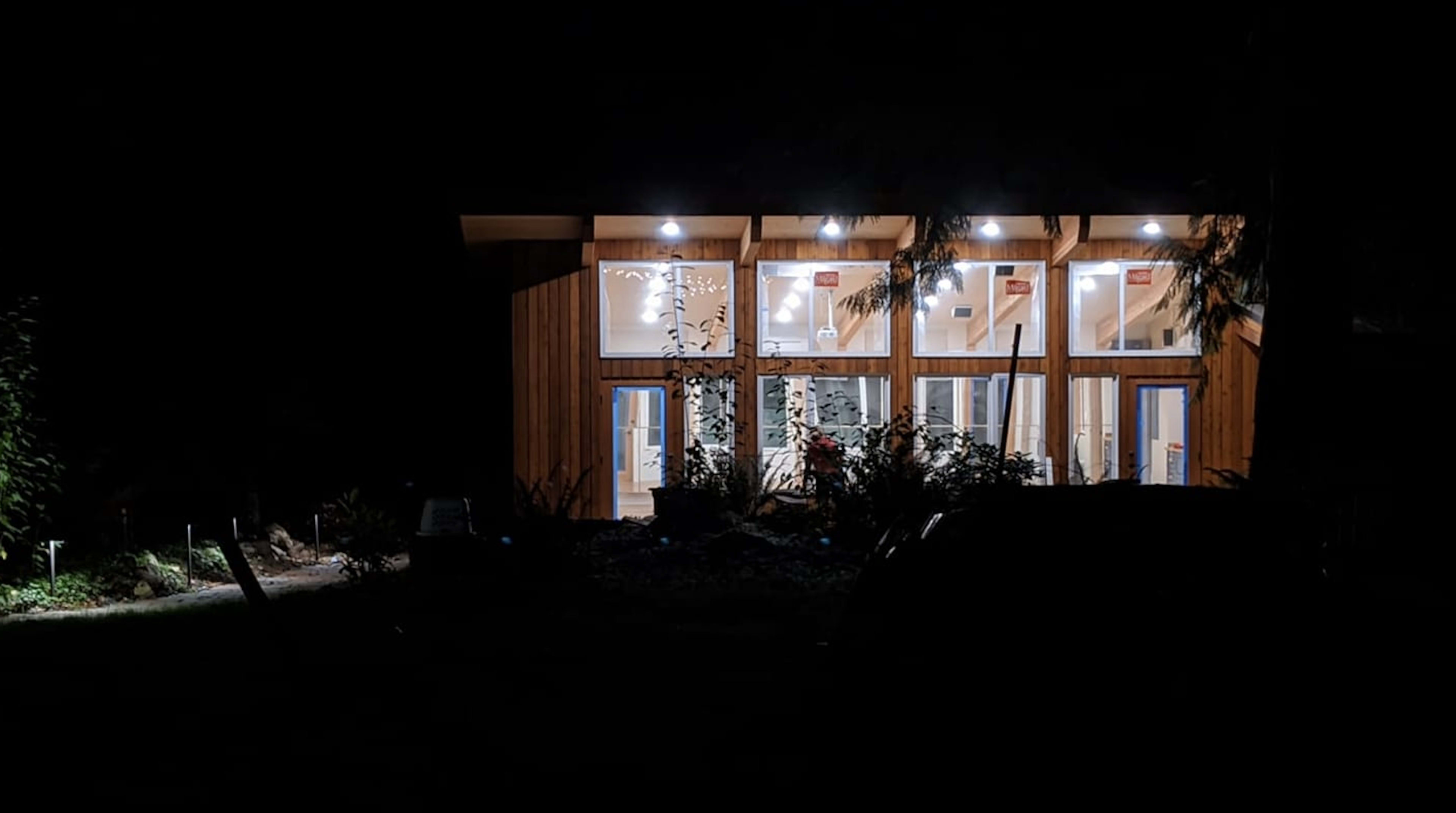A modern wooden structure is illuminated at night, showcasing large glass windows and surrounding greenery.
