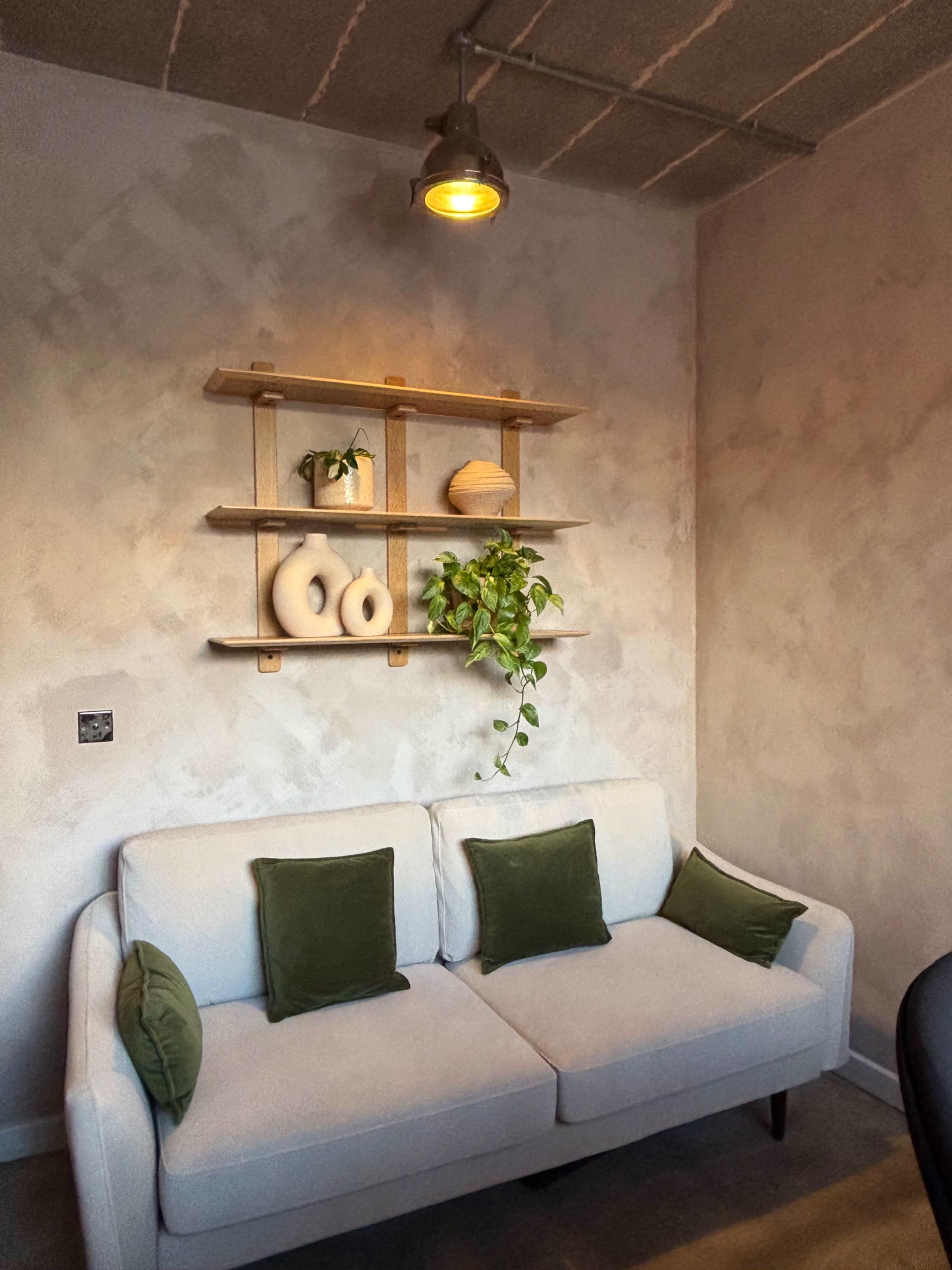The image shows a minimalist living space with a light-colored sofa, two green cushions, and a wall shelf displaying decorative items and a plant.