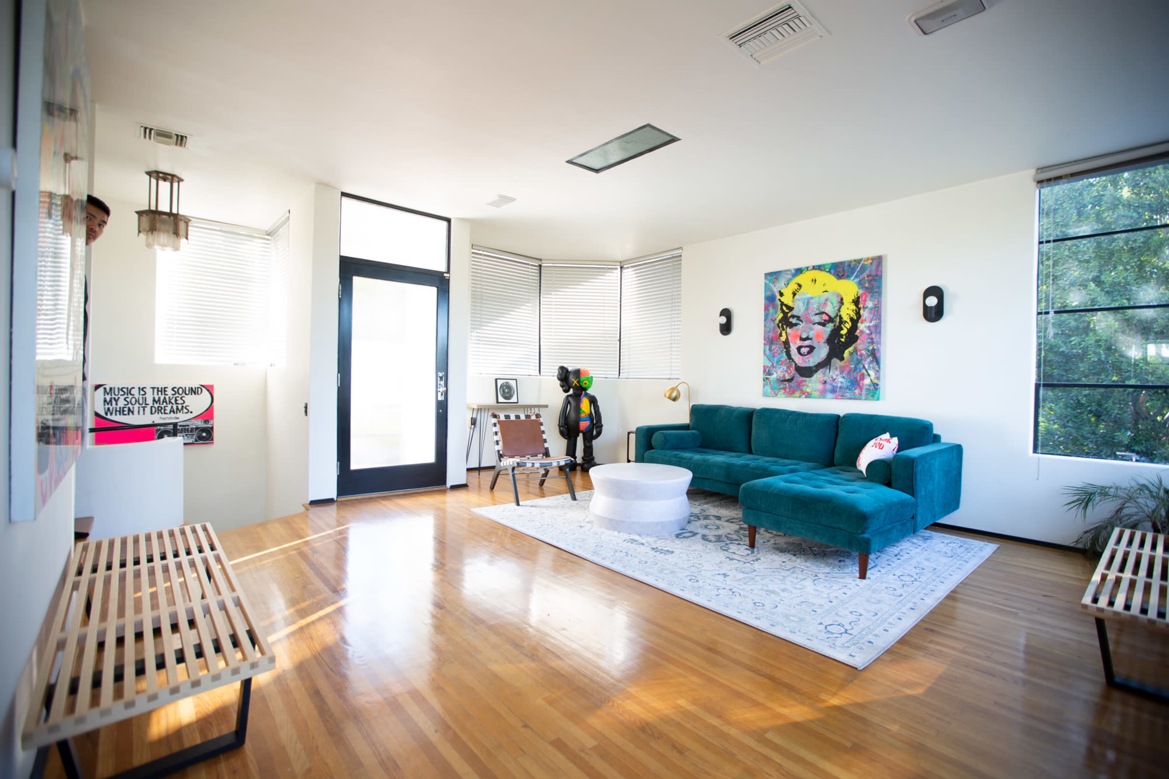 The image shows a modern living room featuring a teal sofa, a circular coffee table, wooden benches, large windows, and colorful wall art.