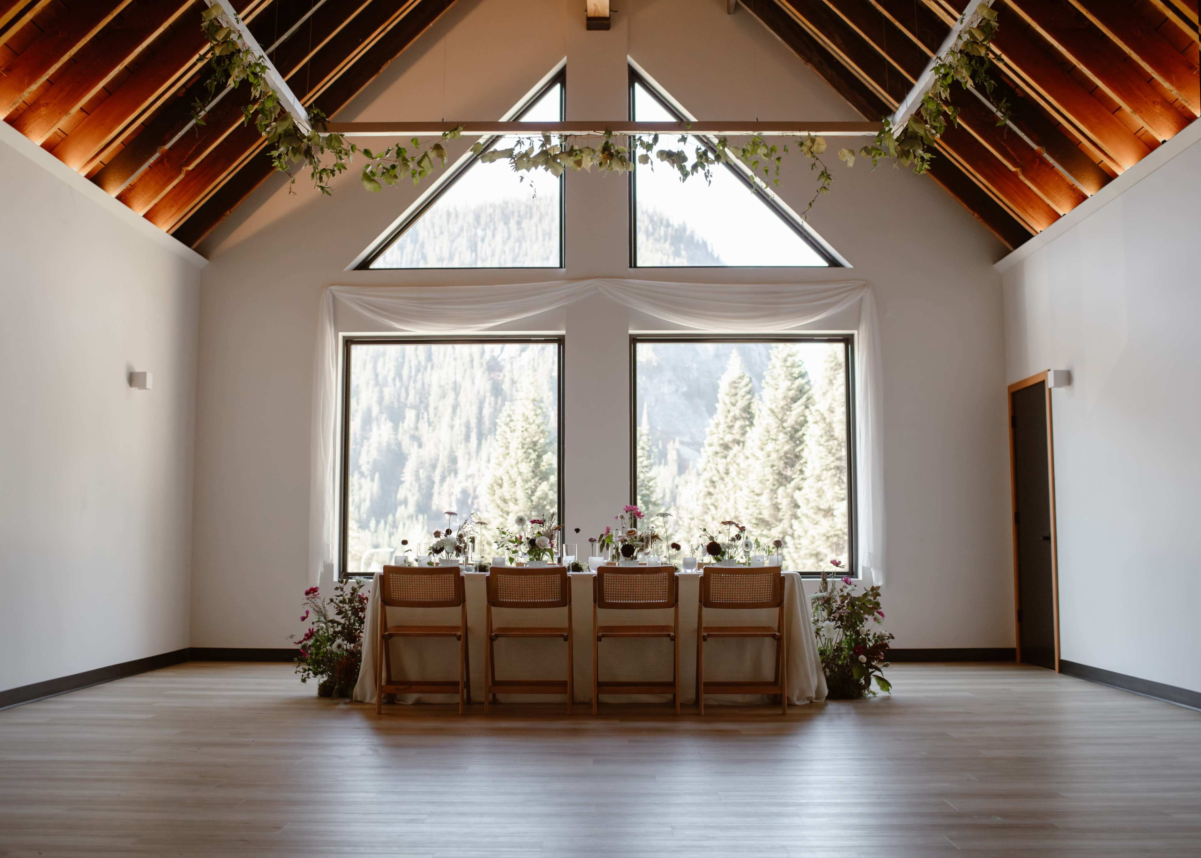 A long dining table with floral arrangements is set against large windows that showcase a forested mountain landscape.