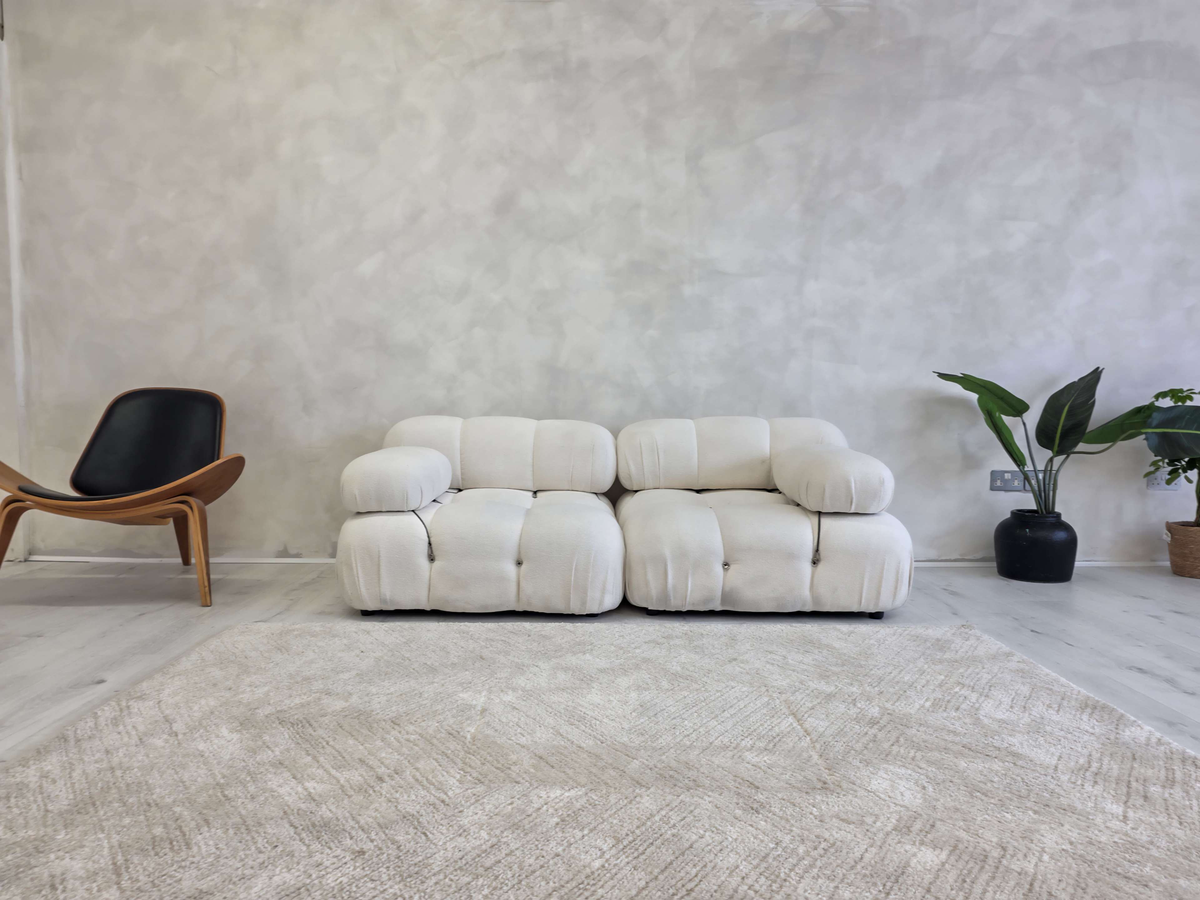 A white, plush two-seater sofa is positioned on a light rug in a minimalist room with a textured wall, a black plant pot, and a wooden chair in the corner.