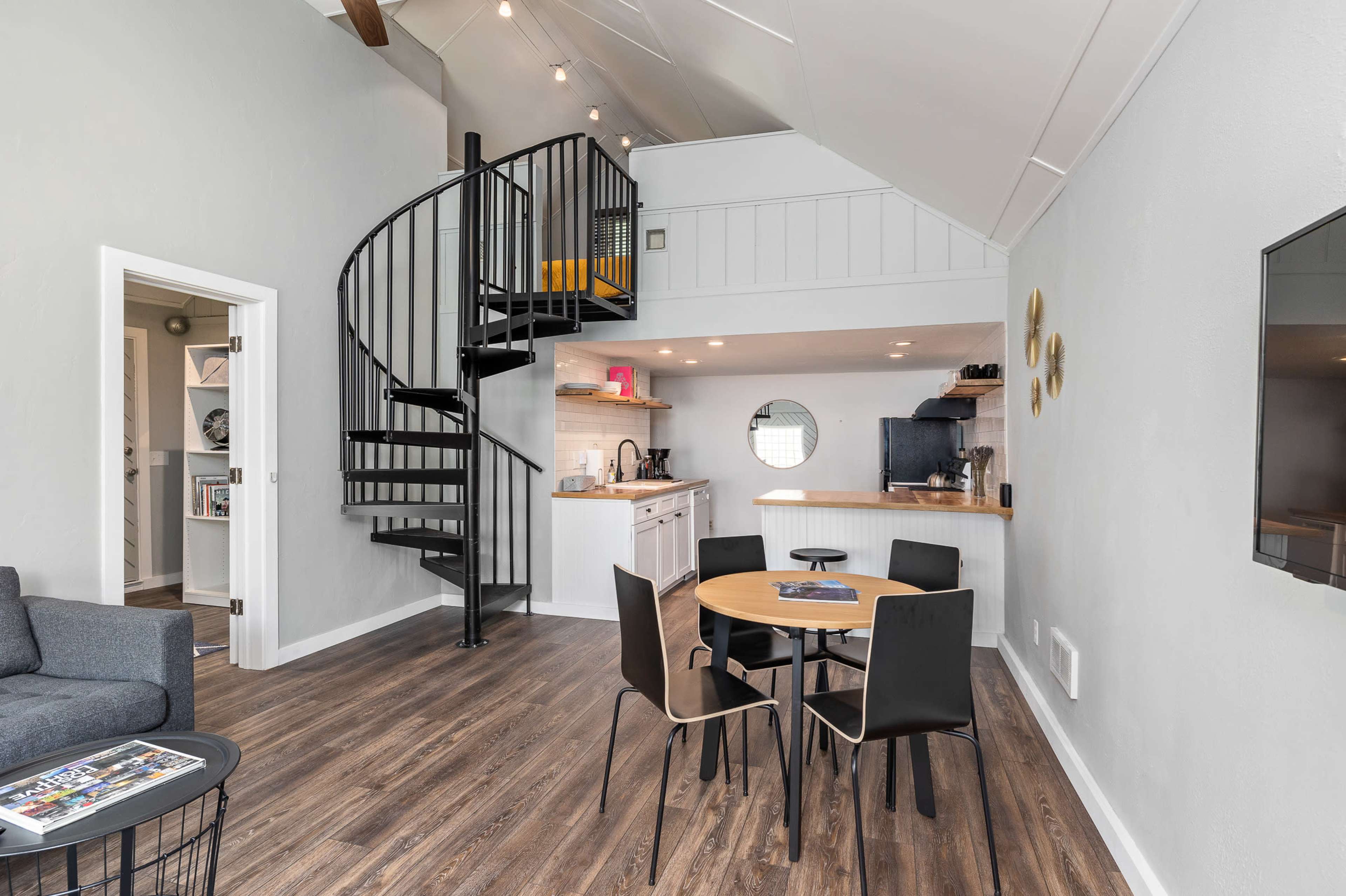 The image shows a modern, open-plan living area with a spiral staircase leading to an upper level, a dining table with four chairs, and a kitchen area.