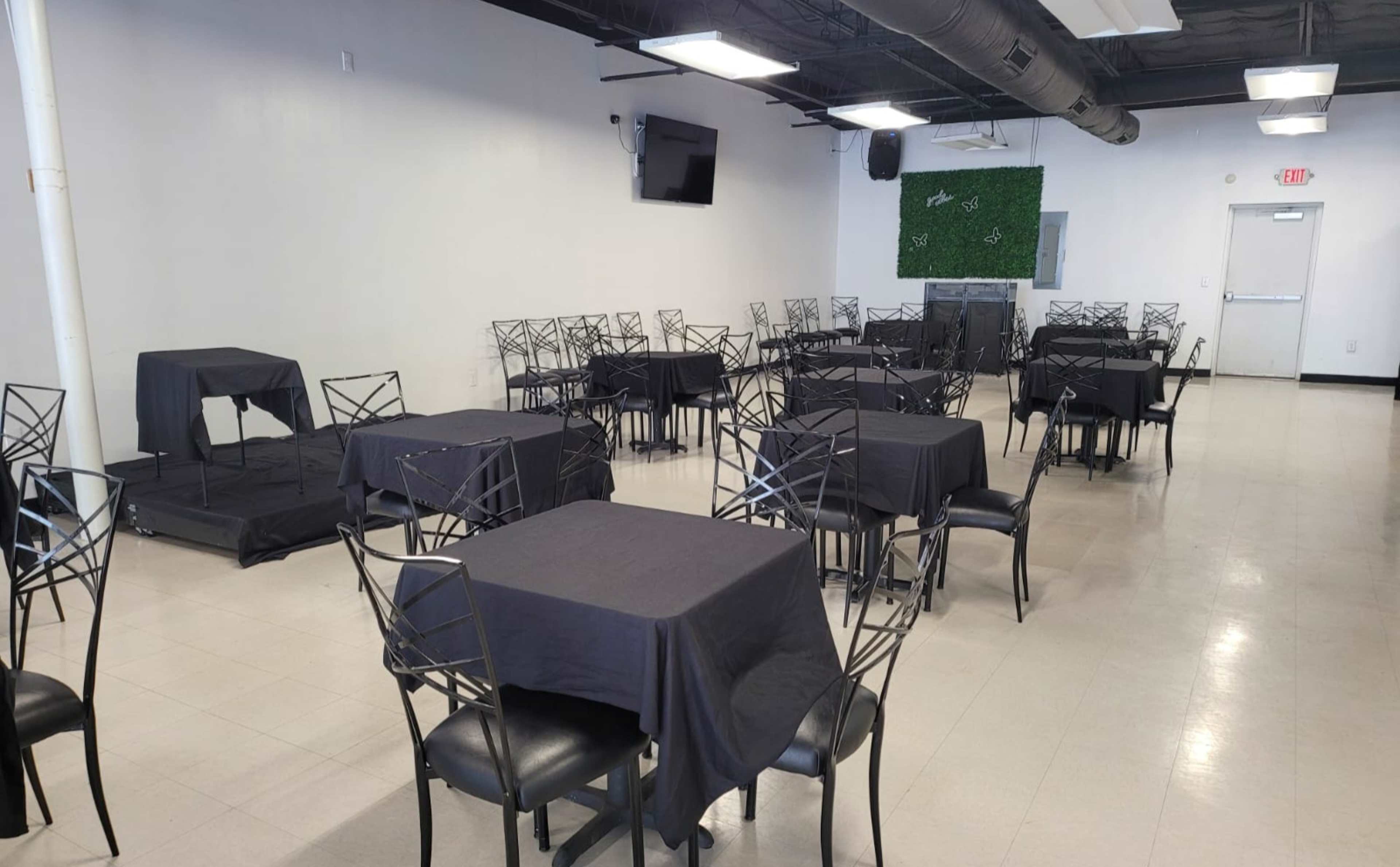 A spacious room features several tables covered with black cloths and metal chairs arranged around them, with a stage and a monitor visible on one side.