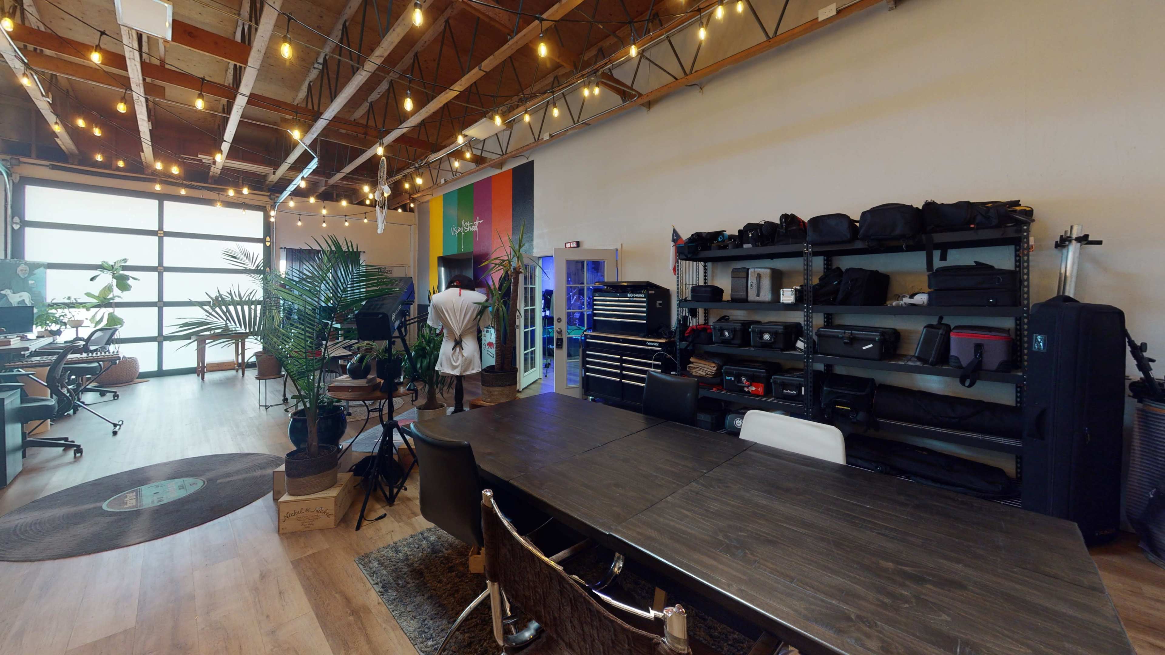 A spacious office-like environment with a large wooden table, chairs, shelves filled with equipment, and plants, illuminated by string lights.
