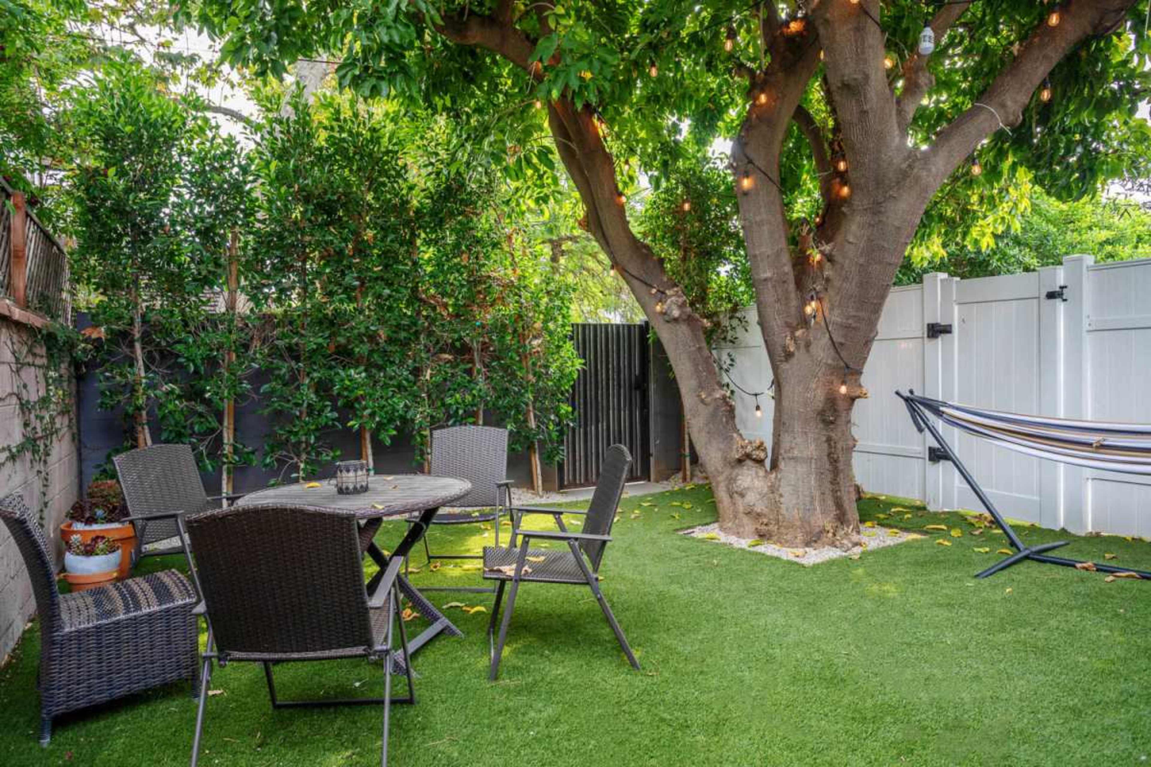 A peaceful backyard featuring a round table with four chairs, a hammock, and a large tree adorned with lights.