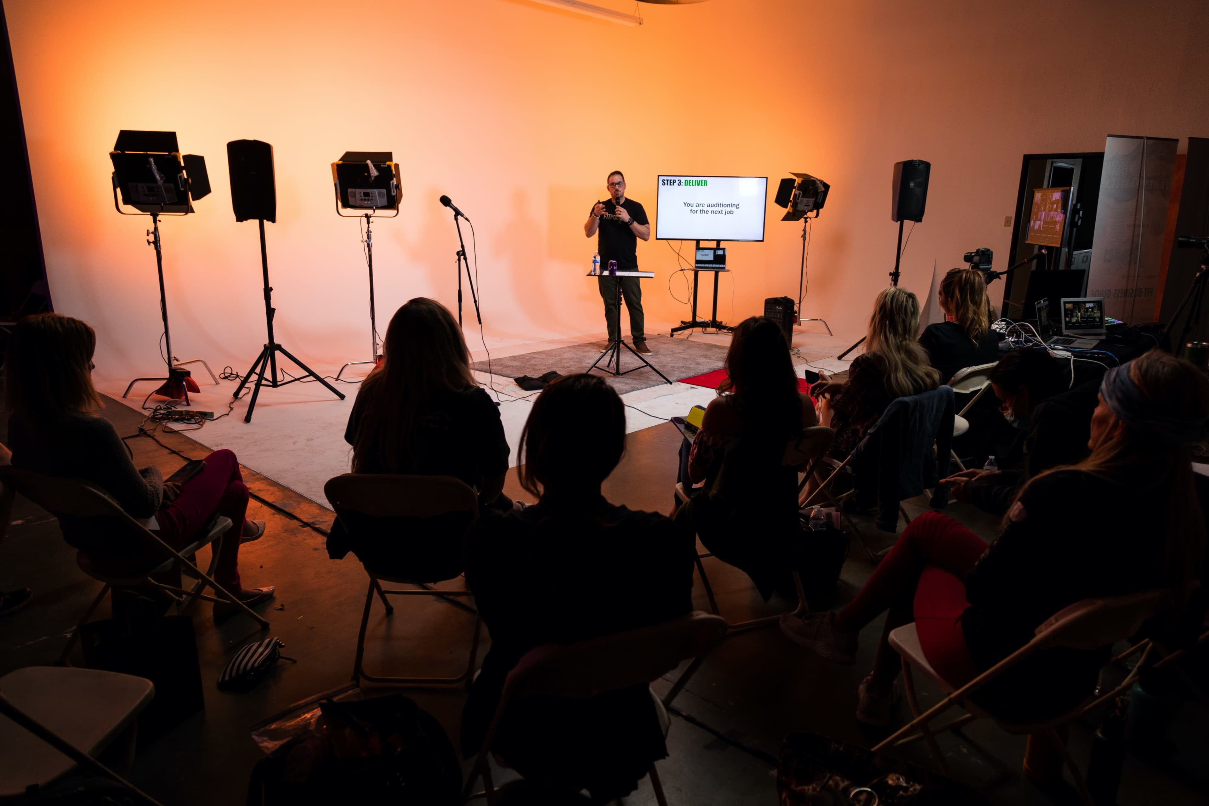 A speaker presents in front of an audience in a studio lit with warm orange lighting.
