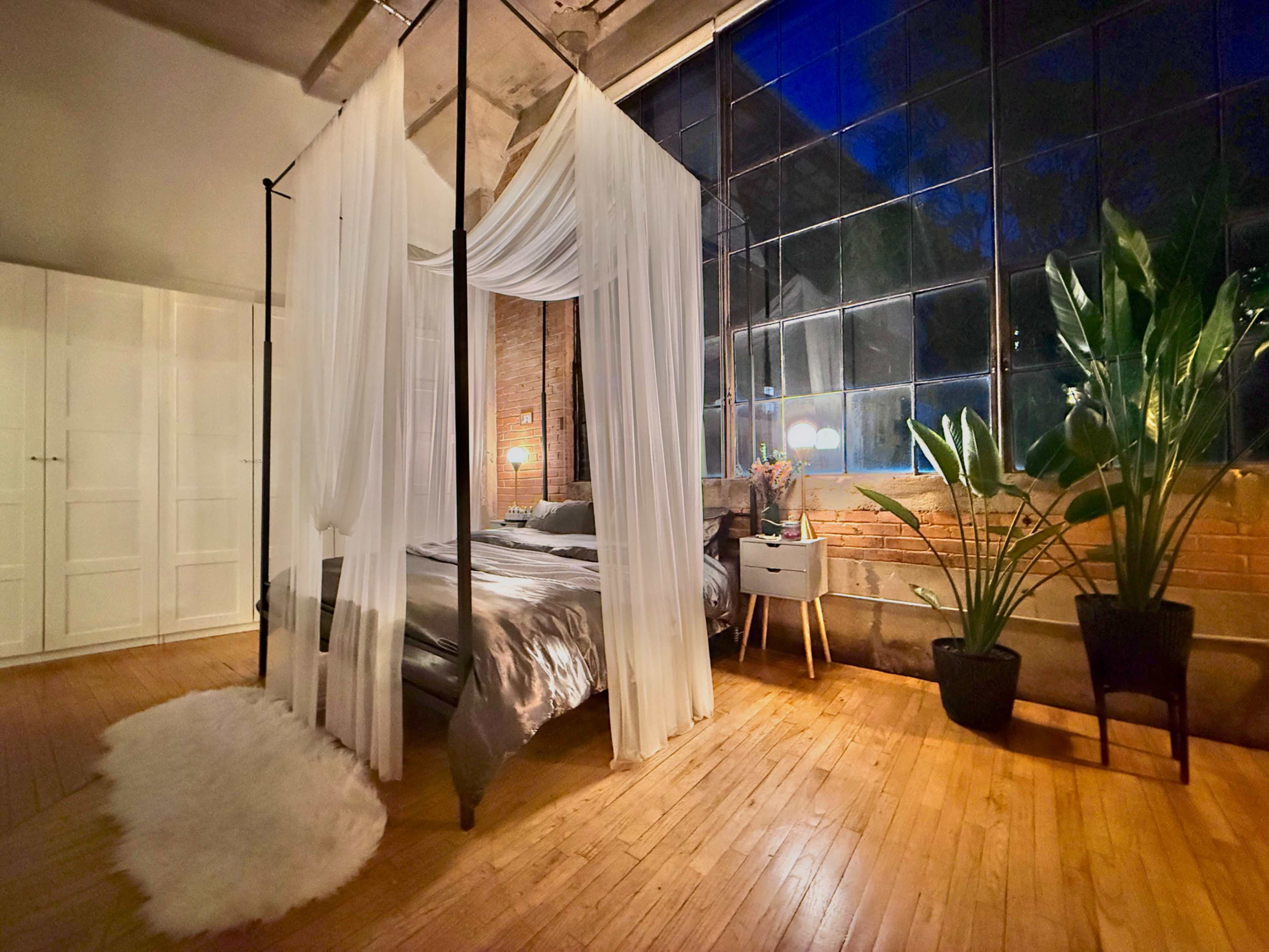 The image shows a bedroom with a four-poster bed draped in sheer curtains, surrounded by large windows and greenery.
