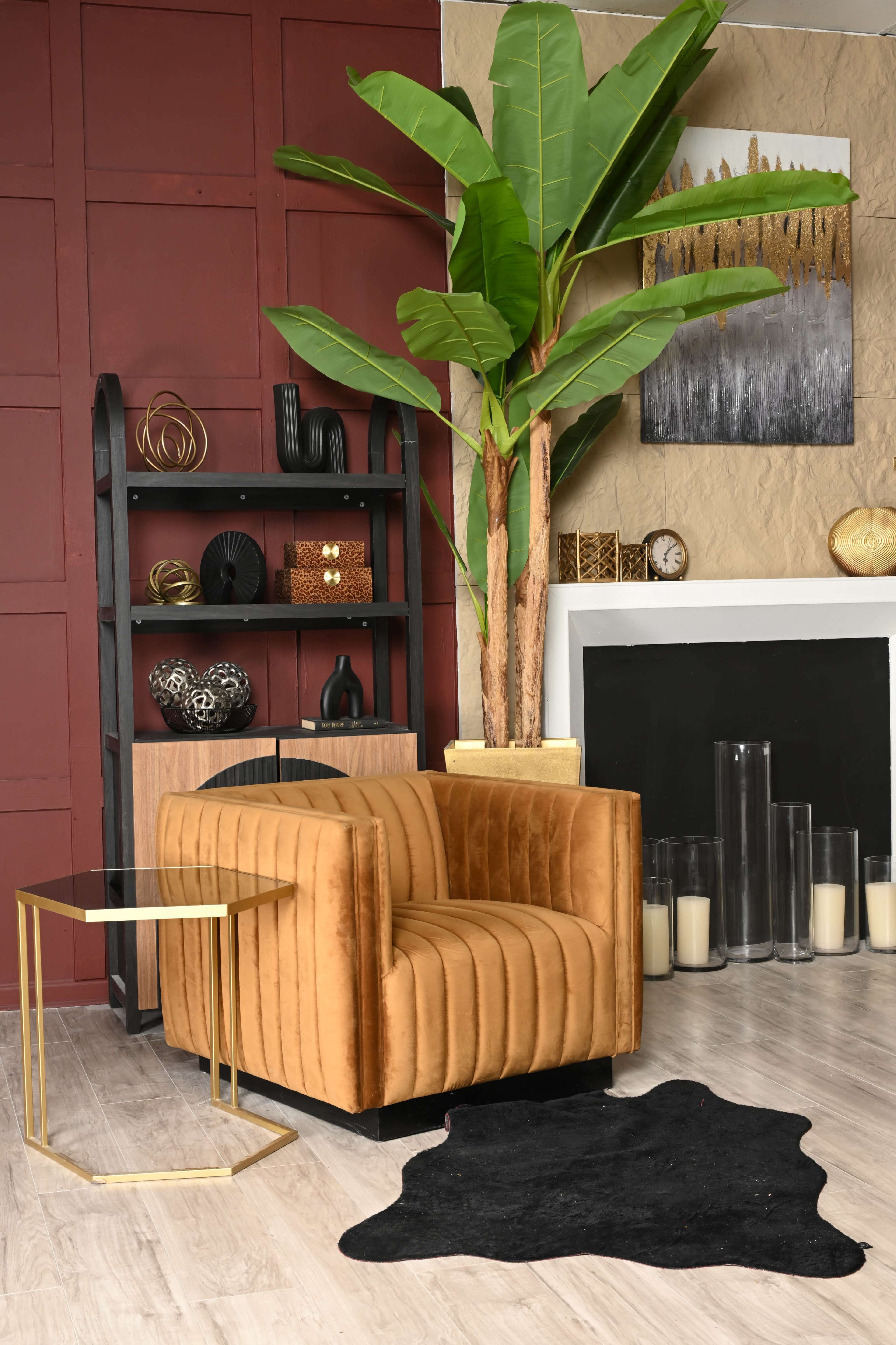 The scene features a stylish interior with a brown velvet armchair, a gold accent table, a large potted plant, and decorative items on a black bookshelf against a maroon wall.