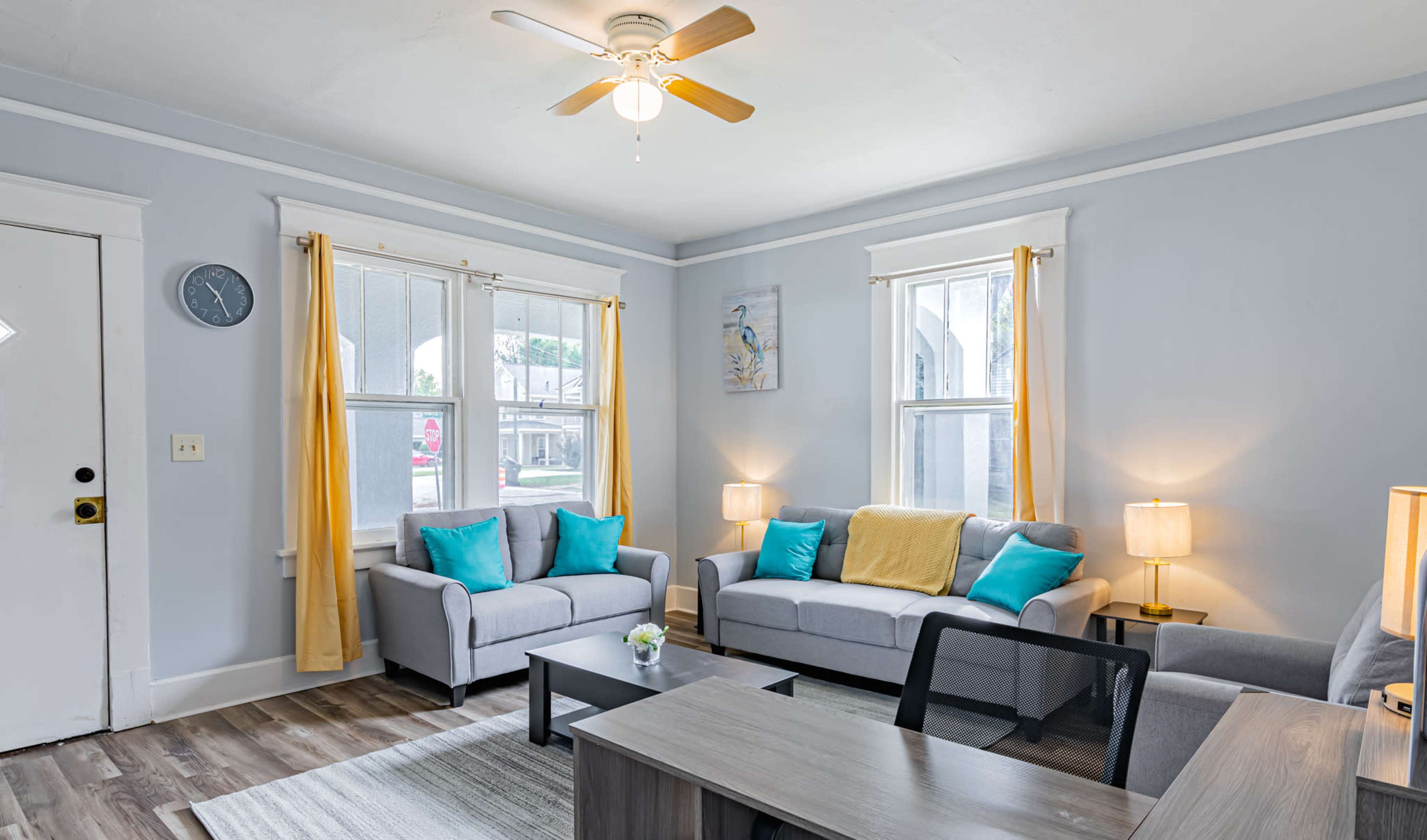 The living room features two gray sofas with blue and yellow accents, a wooden table, and large windows with light curtains.