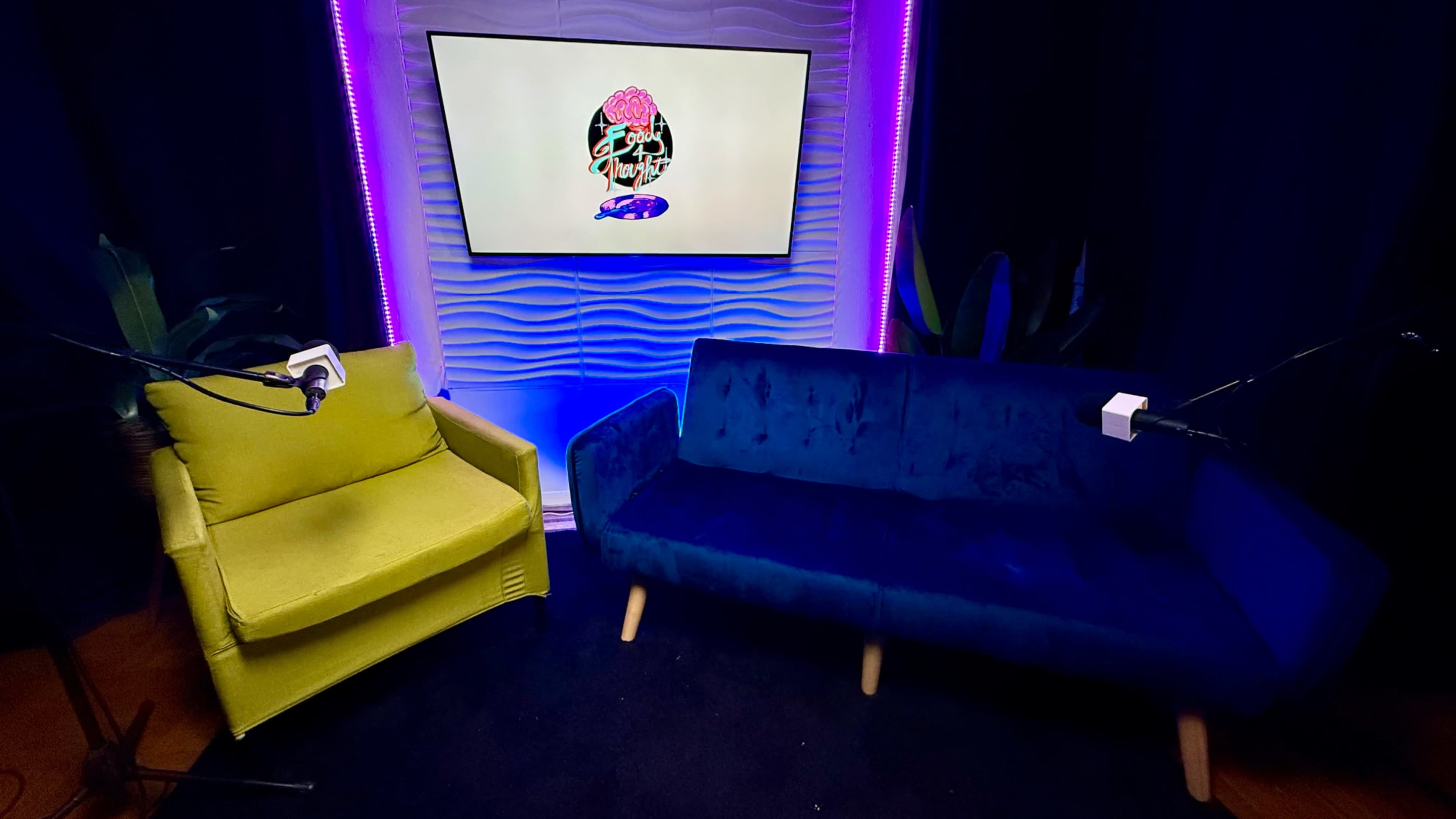 The image shows a recording studio setup featuring a yellow chair and a blue couch, along with a microphone on each seating, against a backdrop of a screen displaying a graphic design.
