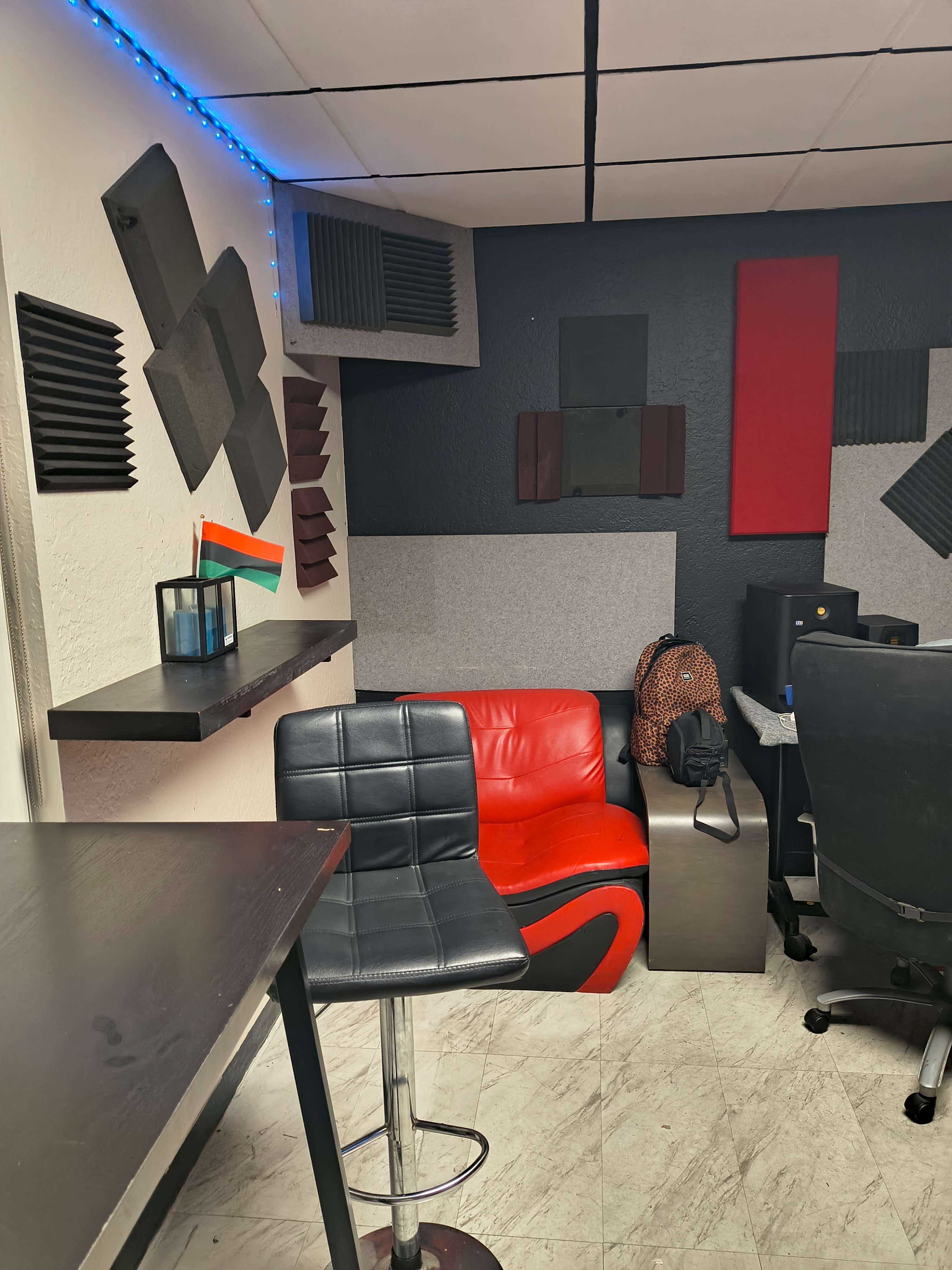 The image shows a small recording studio or workspace featuring soundproofing panels on the walls, a red accent chair, a black bar stool, and various equipment set up on a desk.