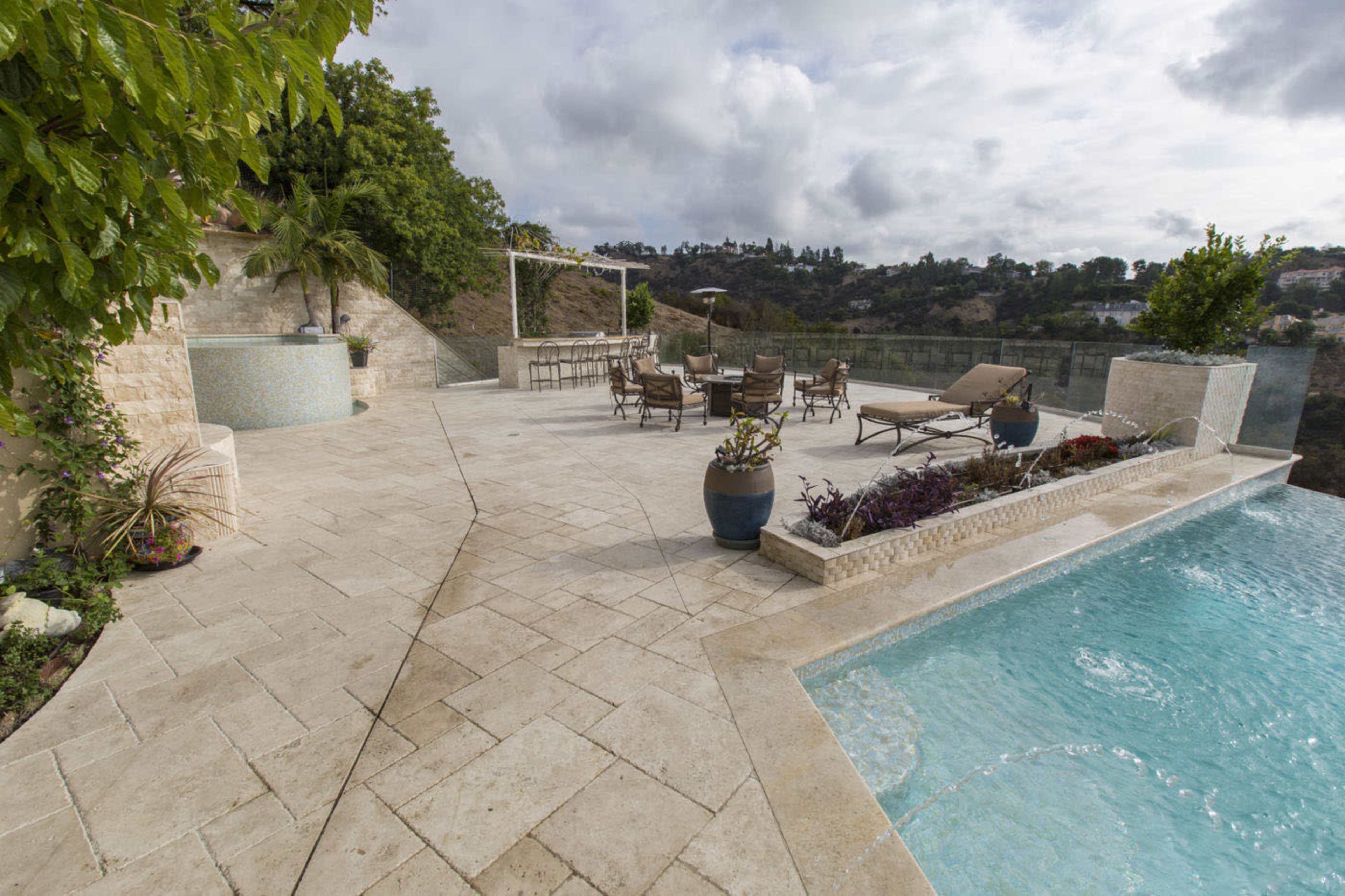 A spacious outdoor patio features a pool, lounge chairs, and a dining area surrounded by greenery and stone walls.