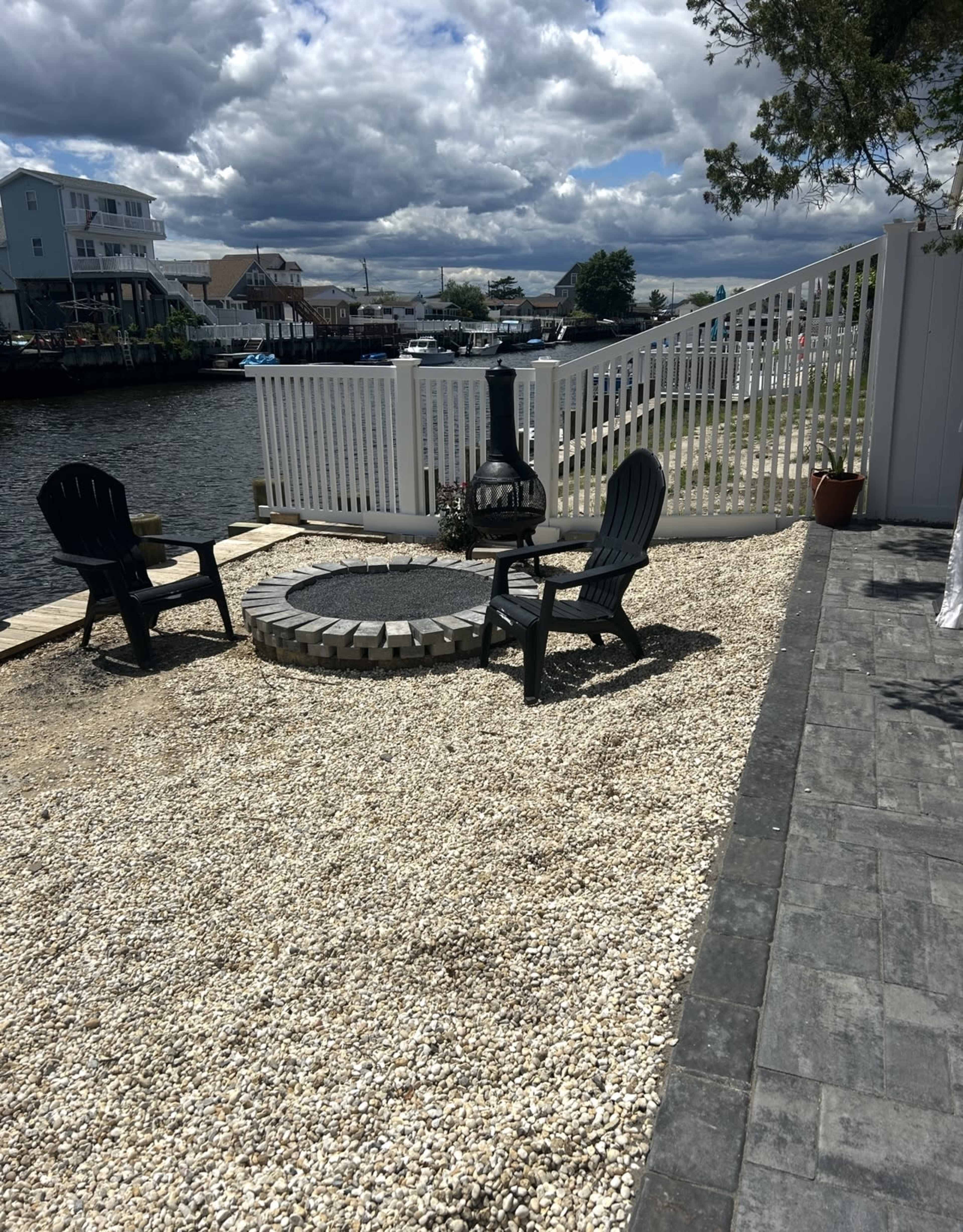 A gravel patio with two black chairs surrounds a circular stone fire pit near a waterway, under a partly cloudy sky.
