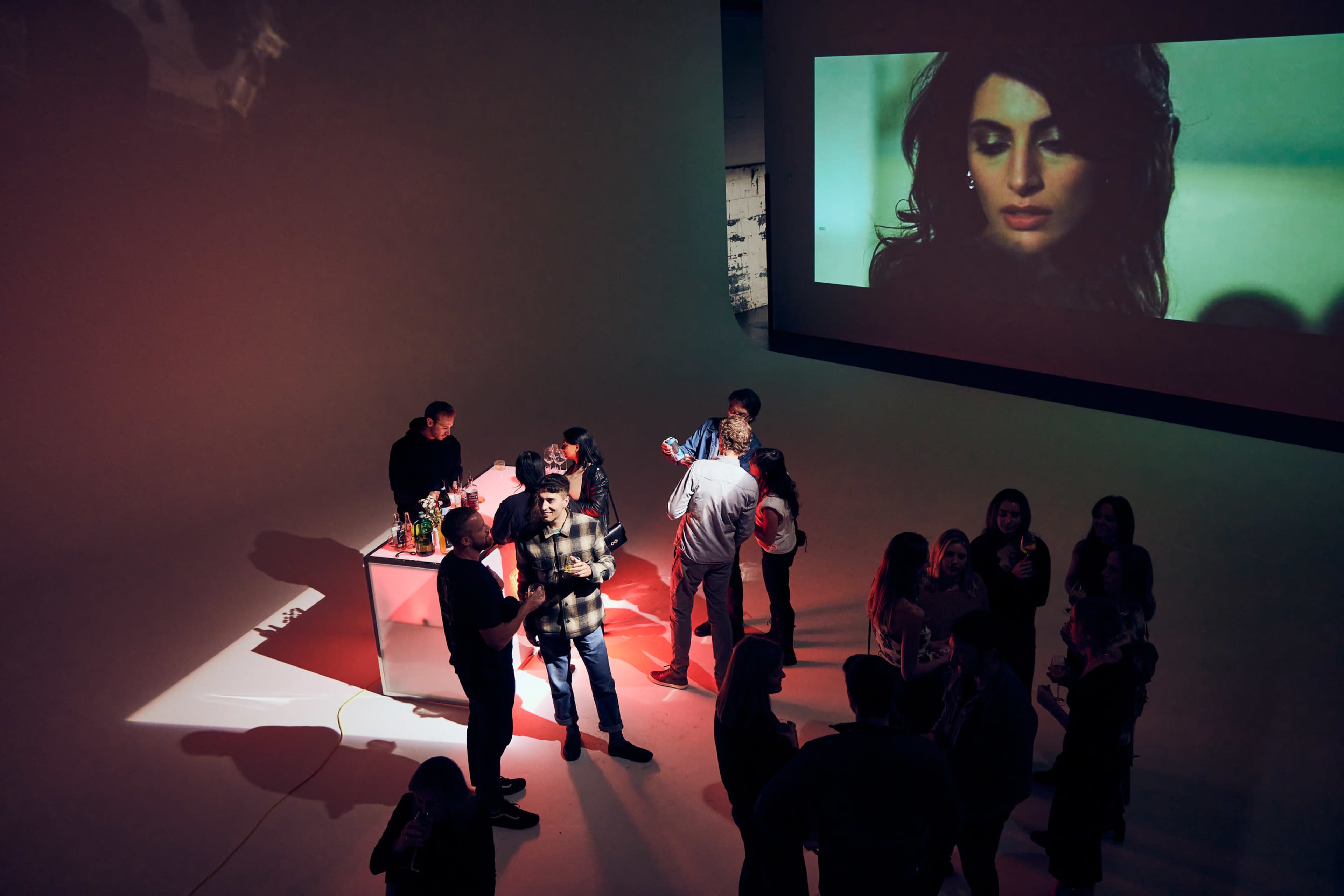 A group of people gathers in a dimly lit space with a large screen displaying a woman's face in the background.
