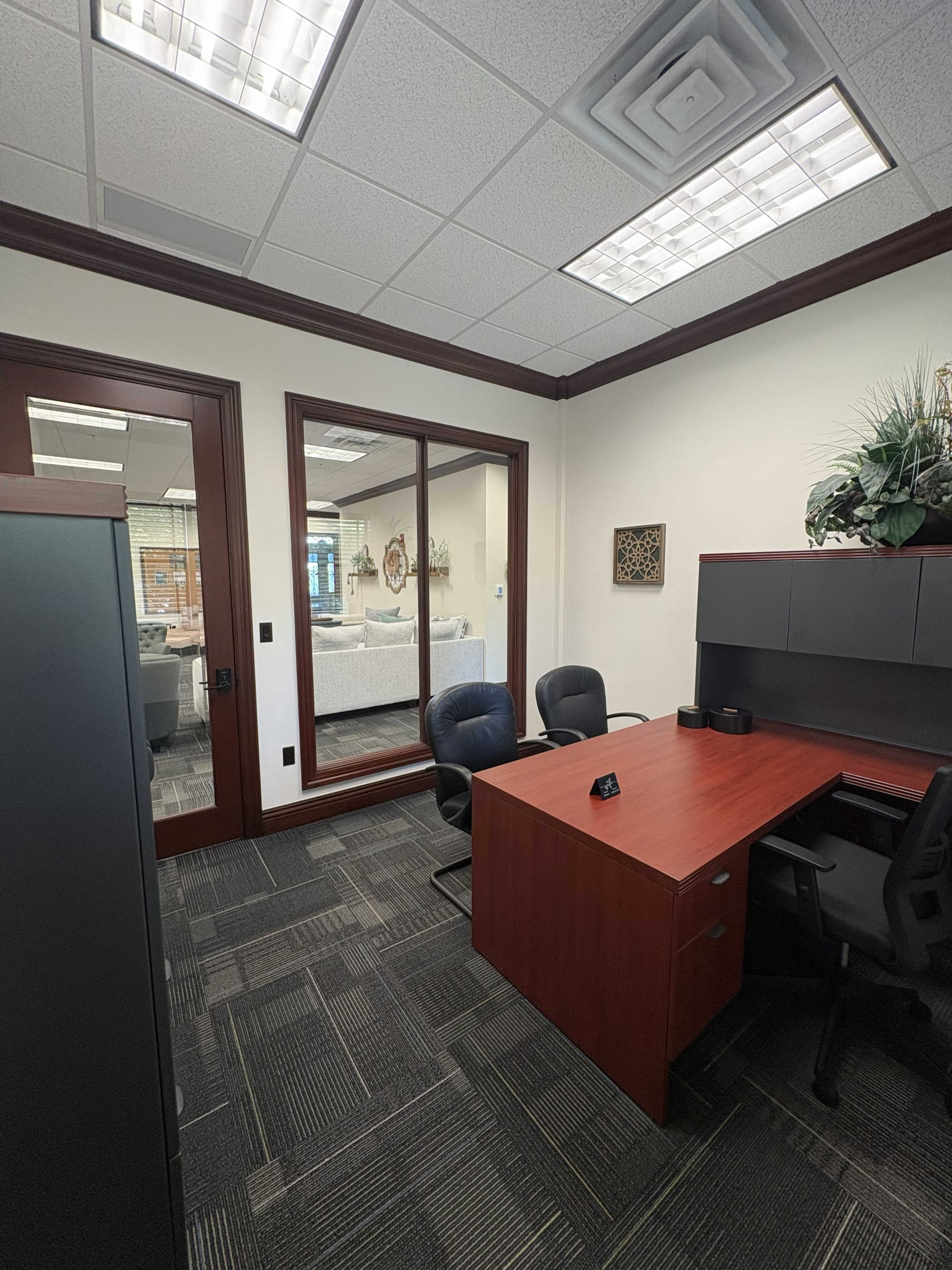 The image shows a modern office space featuring a desk with two chairs, a filing cabinet, and a glass partition revealing a separate seating area.