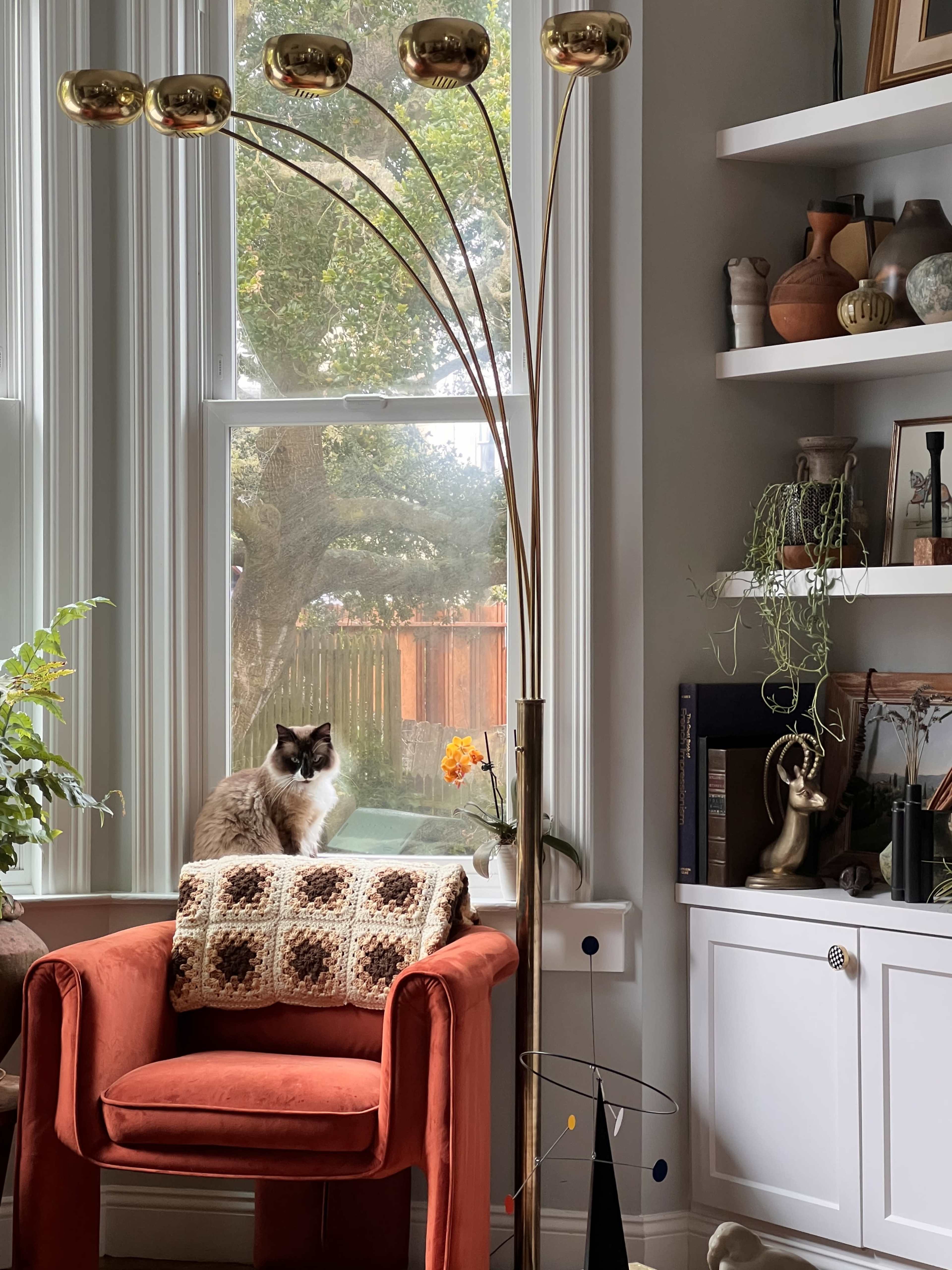 A cat is sitting on a windowsill next to an orange armchair with a patterned blanket, while a standing lamp and various decorative items are placed on a nearby shelf.