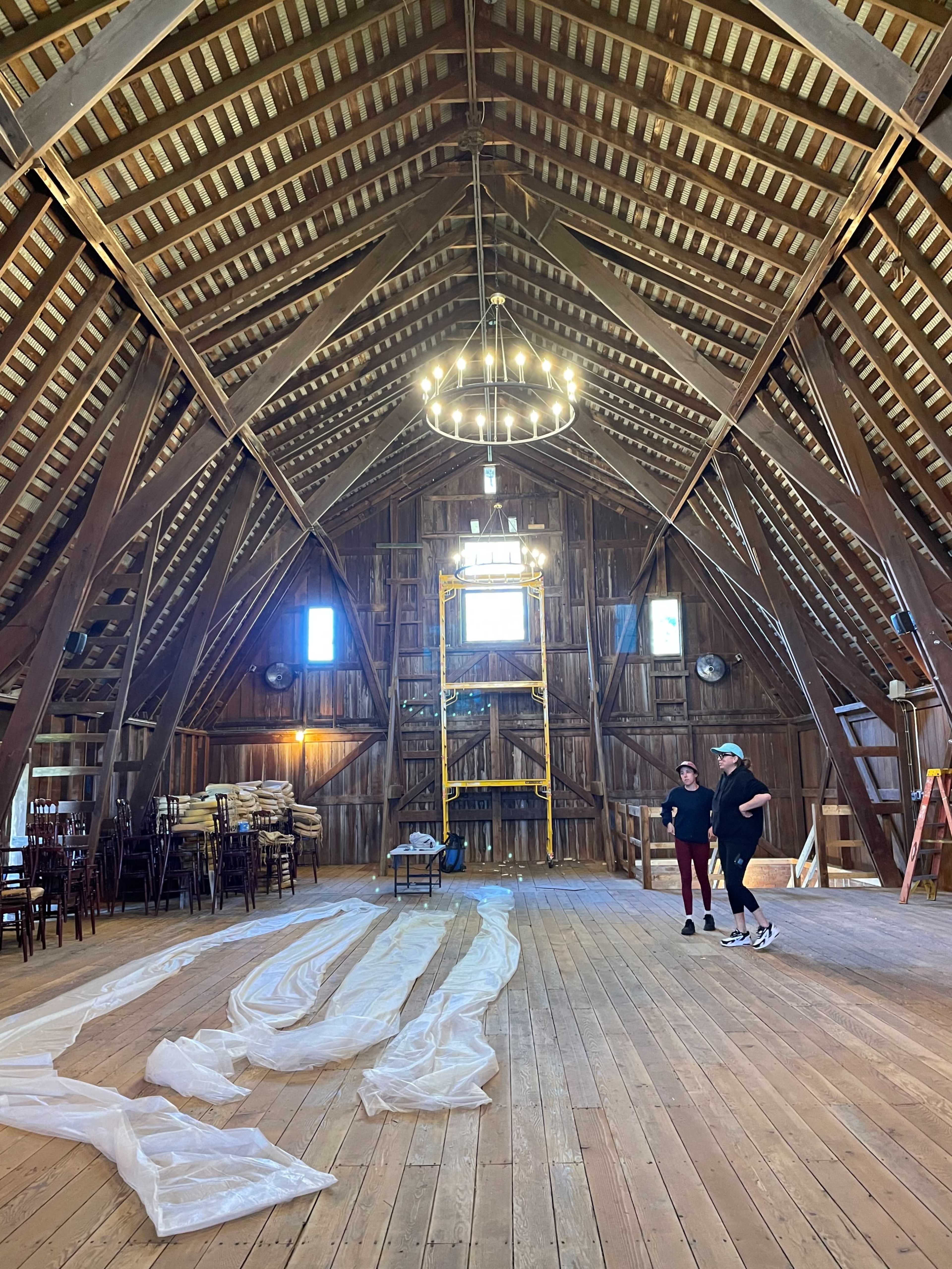 Two people stand in a spacious barn with high, wooden beams and a wooden floor, while a chandelier hangs from the ceiling and white fabric is spread across the floor.