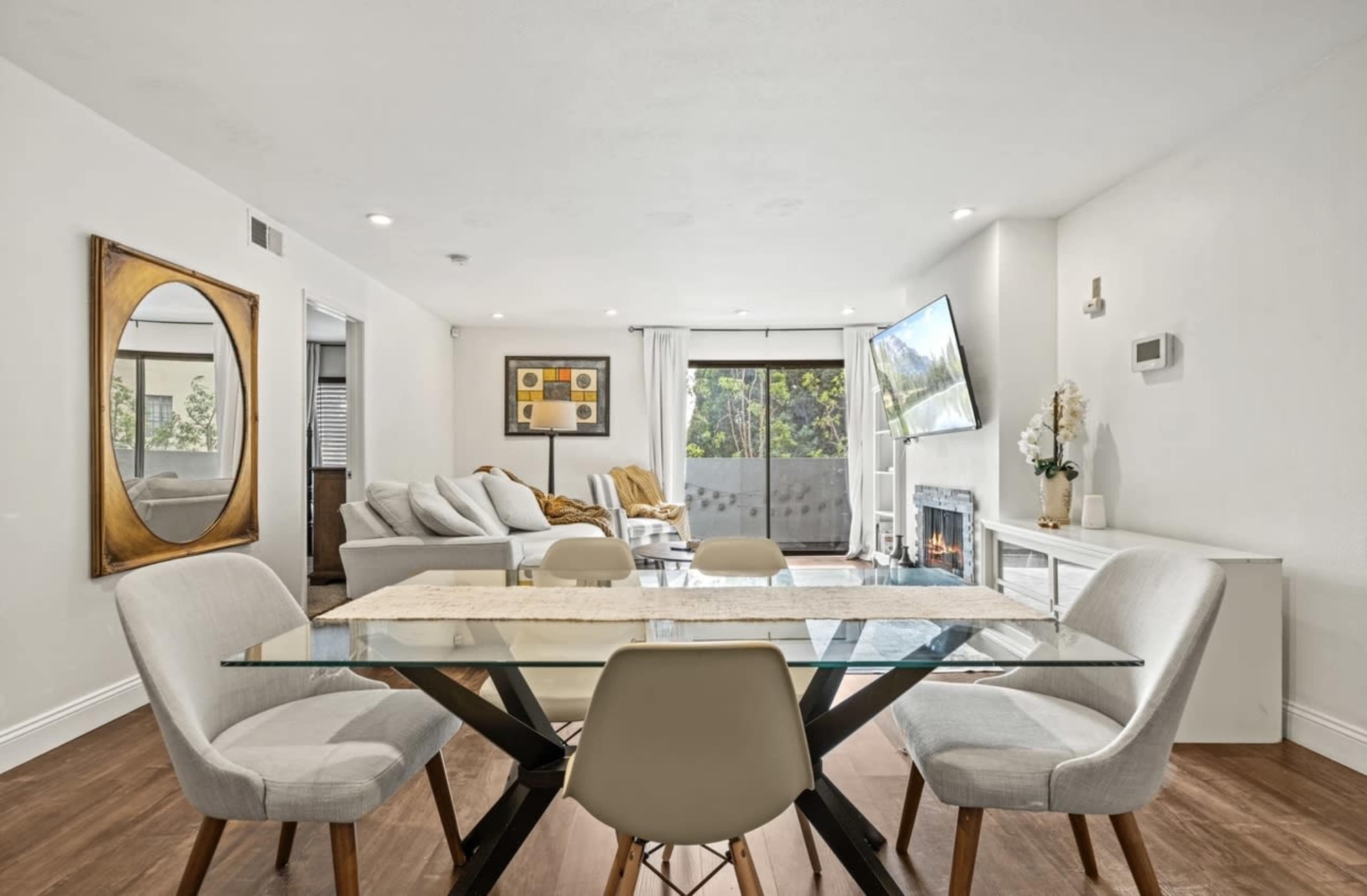 The image shows a modern living area with a glass dining table, light-colored furniture, and a large window overlooking a balcony.