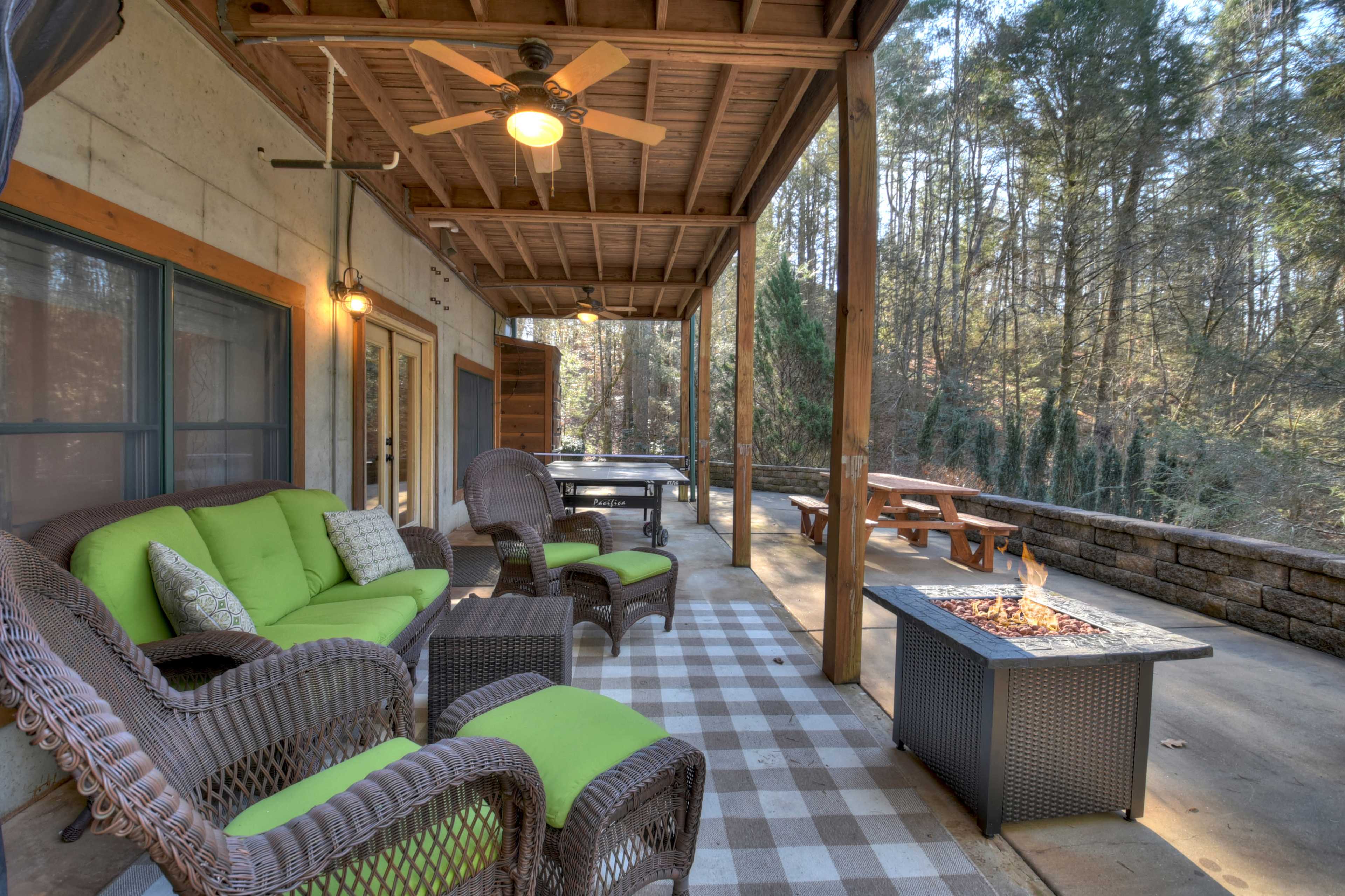 A covered patio with wicker furniture, a fire pit, and a picnic table, surrounded by trees.