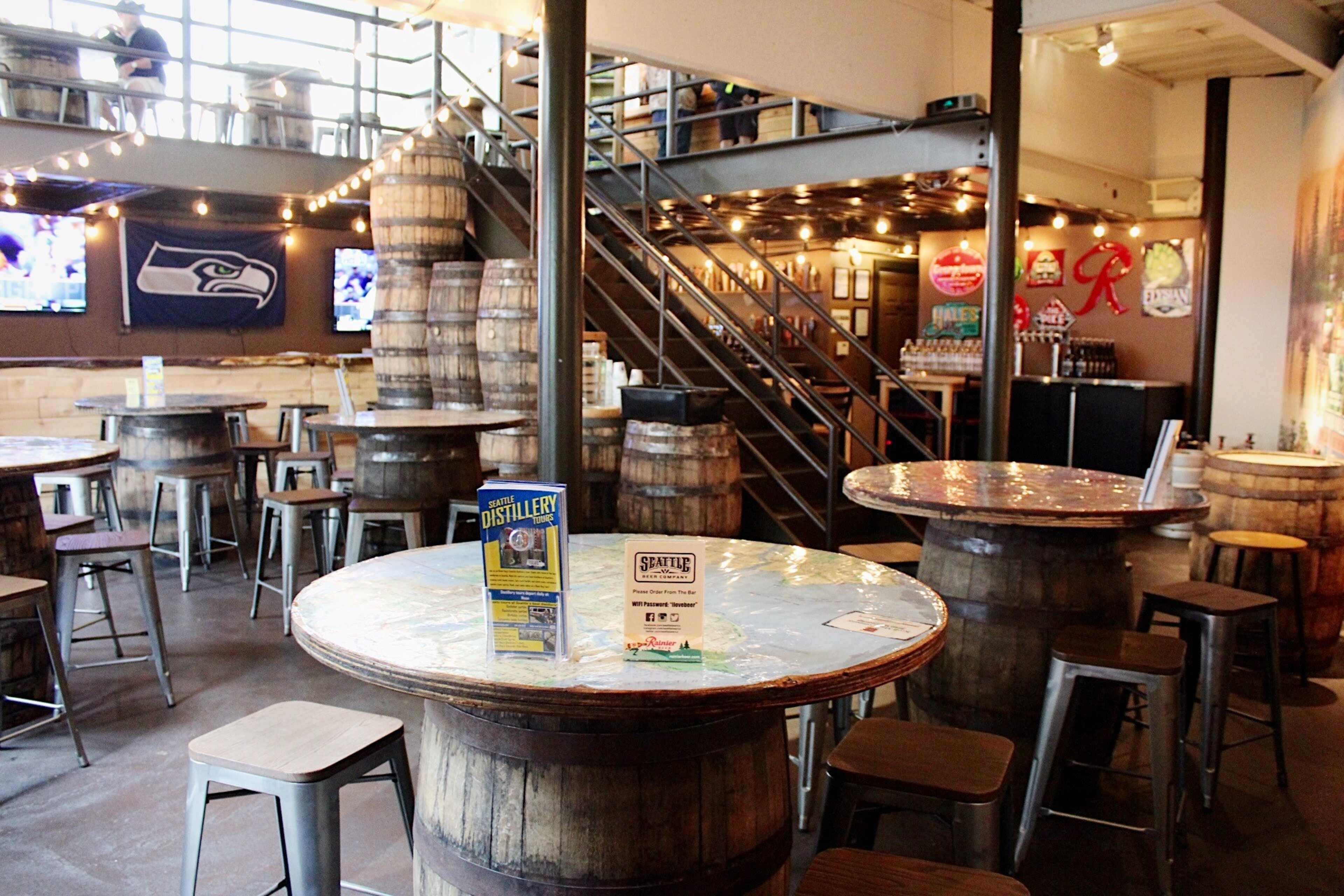 A casual bar setting with wooden barrel tables, metal stools, and a staircase, featuring shelving with drinks and a TV displaying a sports event.