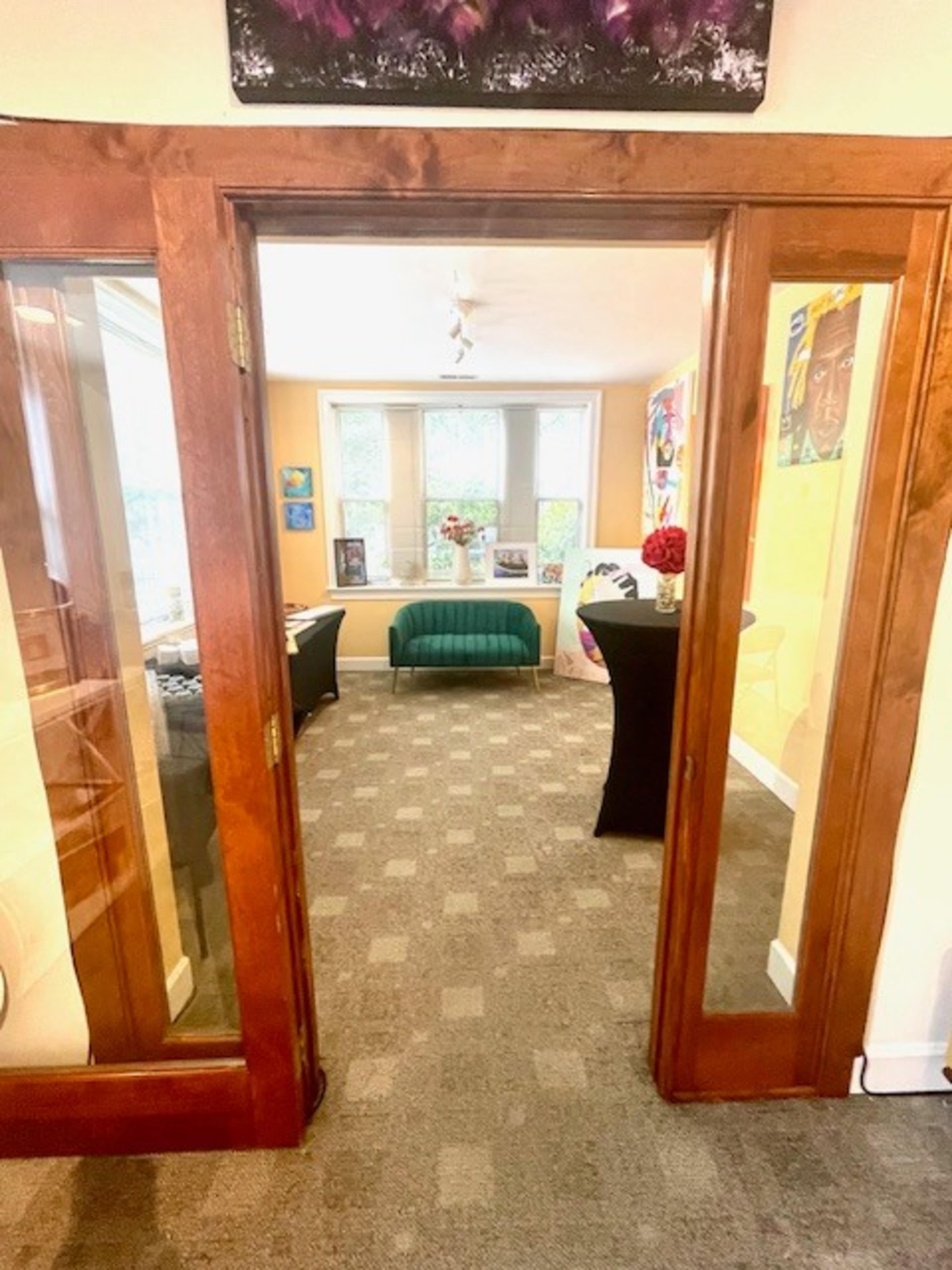 The image shows a view through two wooden doors into a brightly lit room featuring a green sofa, art on the walls, and a carpeted floor.