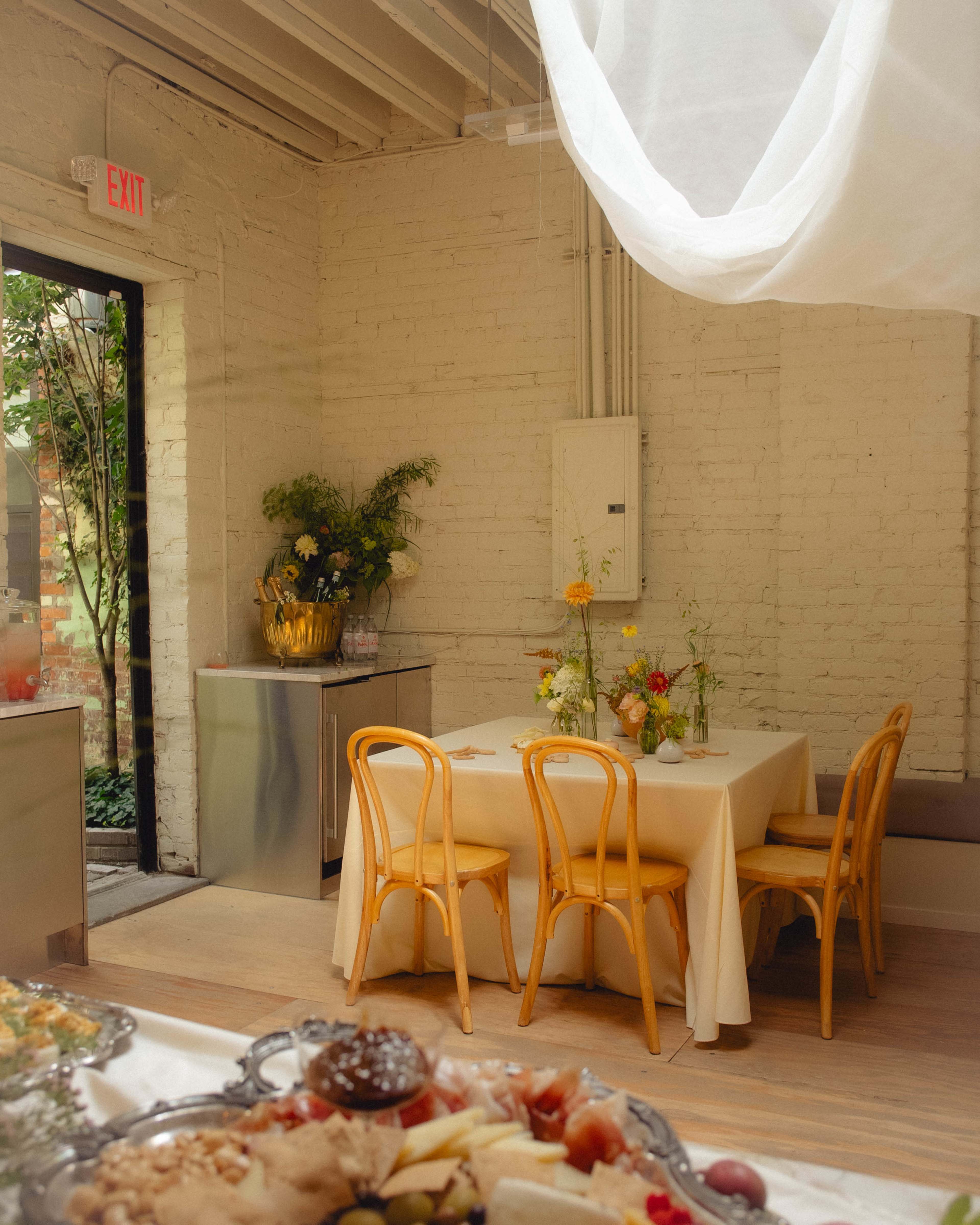 A small dining area features a table with floral centerpieces and yellow chairs next to a large window with greenery outside.