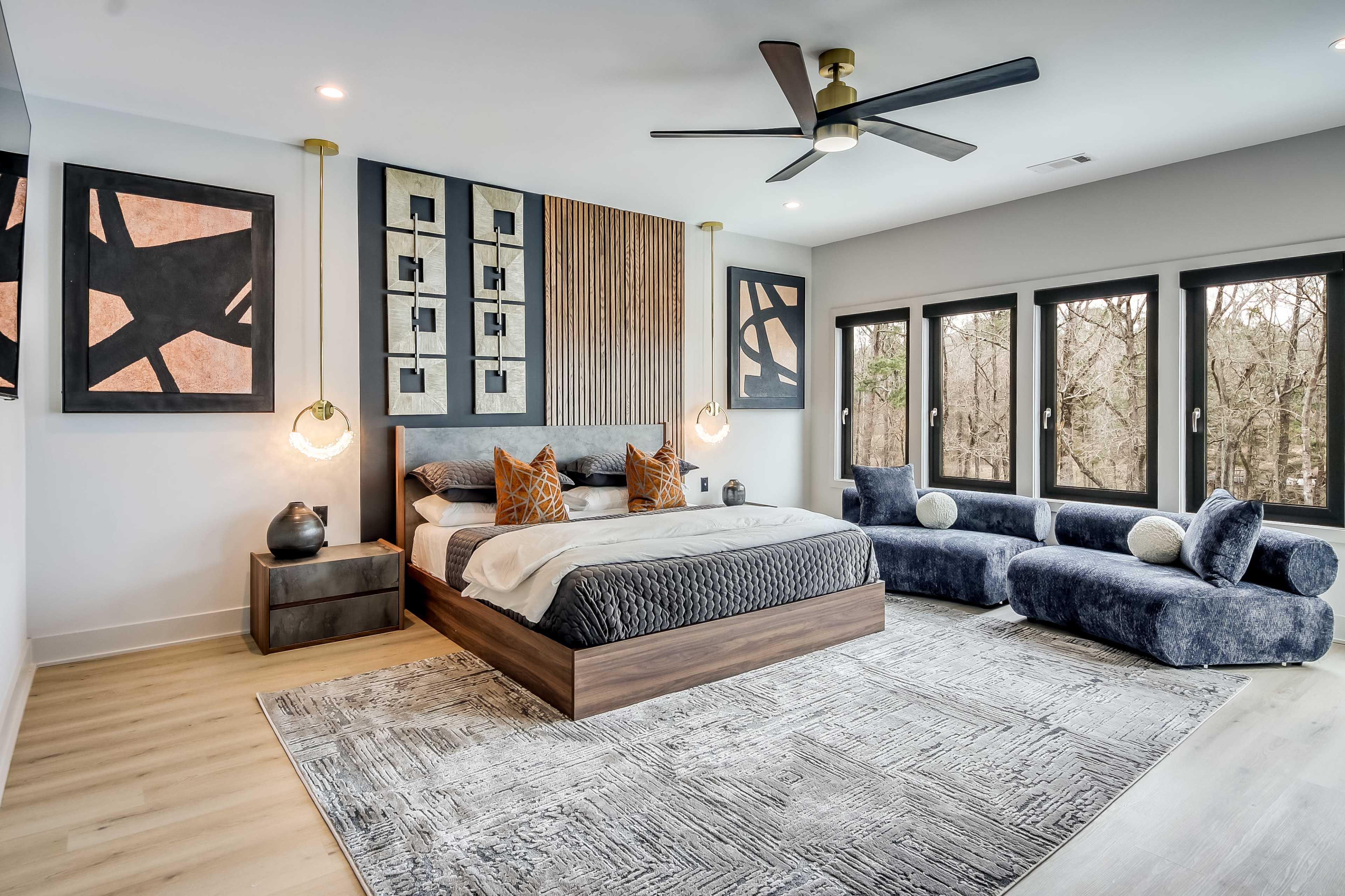 A spacious modern bedroom features a large bed with decorative pillows, a blue sectional sofa, and large windows overlooking a wooded area.