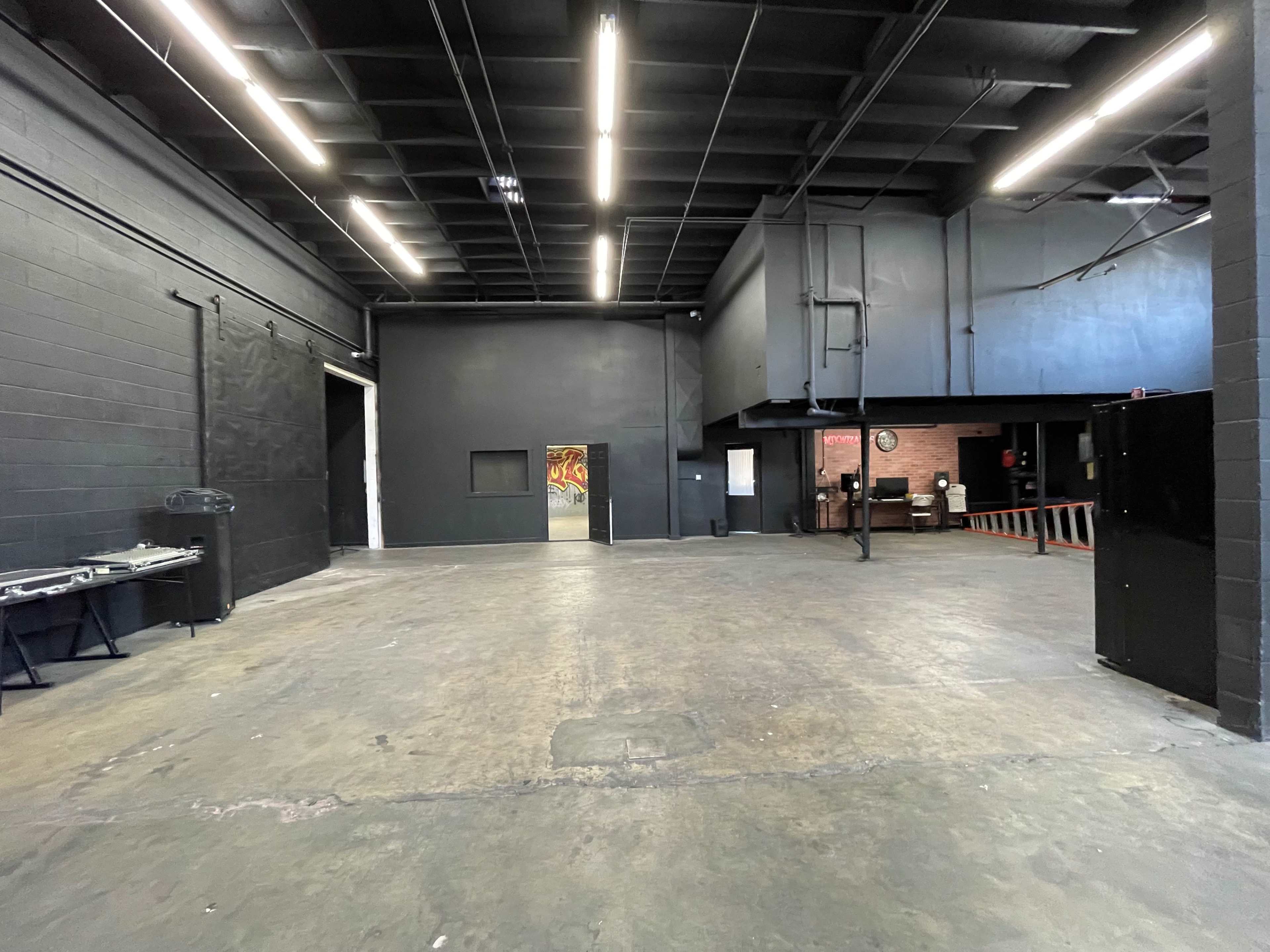 A spacious, empty indoor venue with black walls and a concrete floor, featuring a loading door and a small stage area in the background.