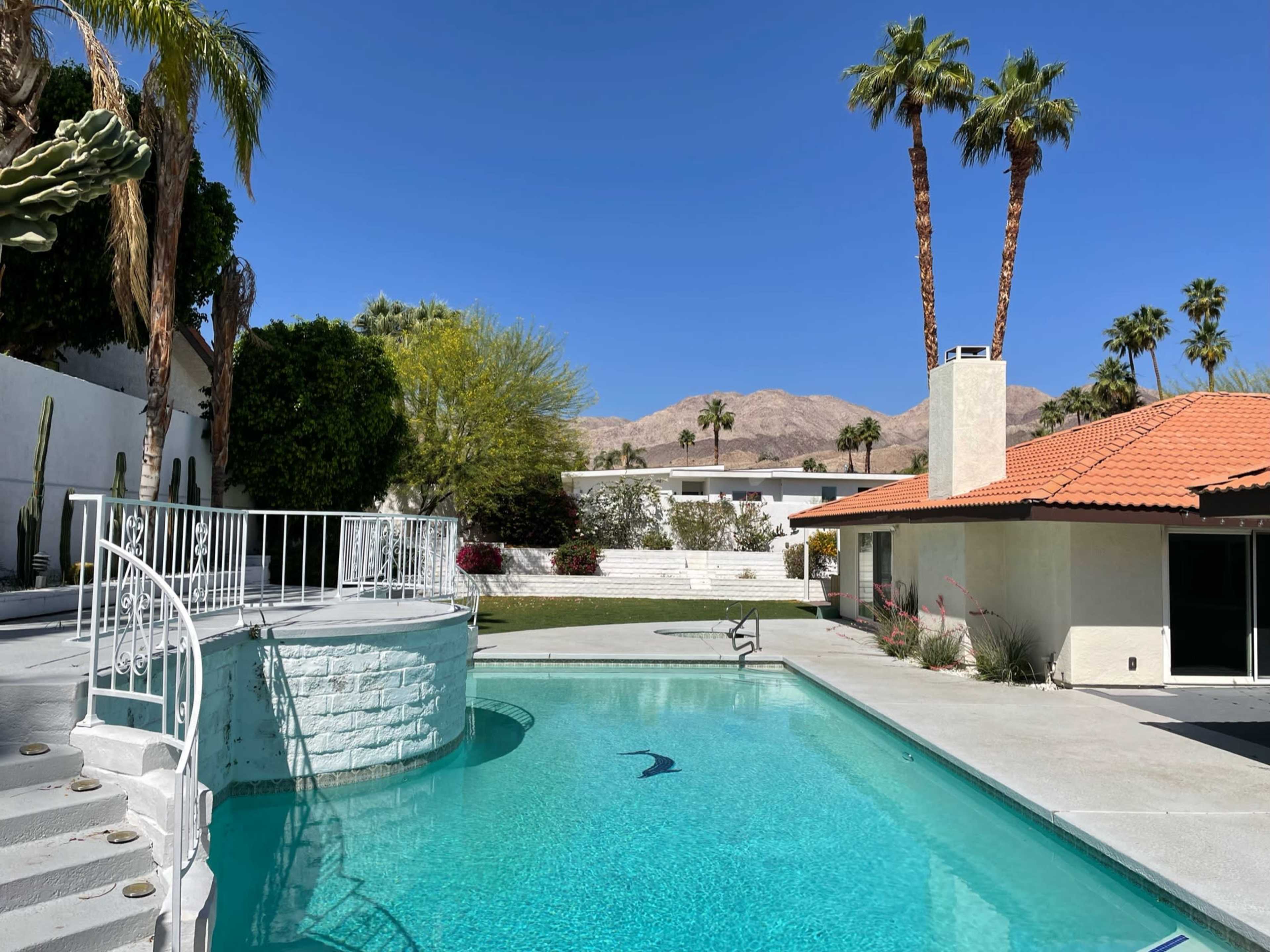 A swimming pool is situated in a landscaped backyard, surrounded by palm trees and mountains in the background.