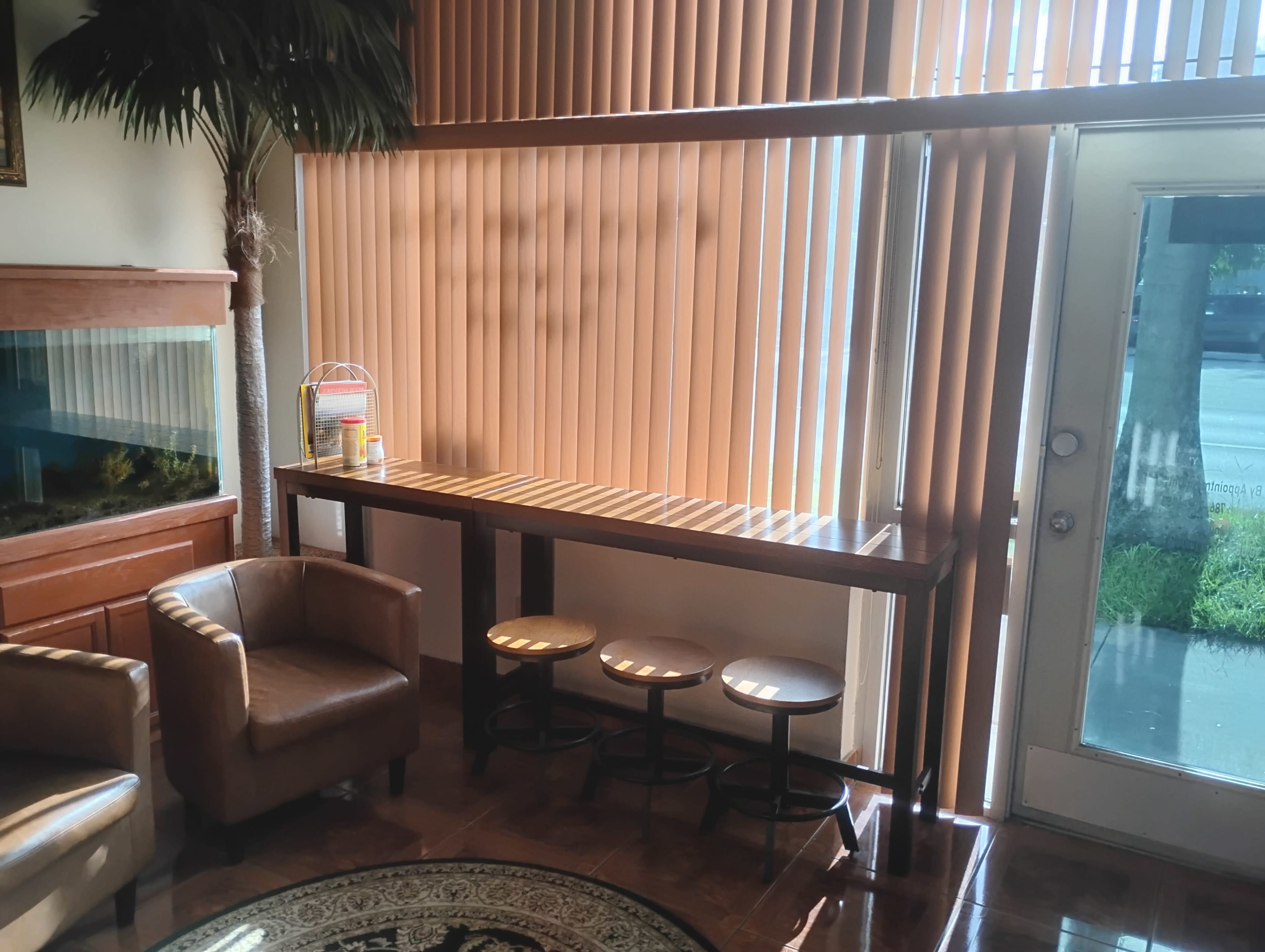 A wooden table with three stools is positioned near a window with vertical blinds, alongside a fish tank and a brown armchair.