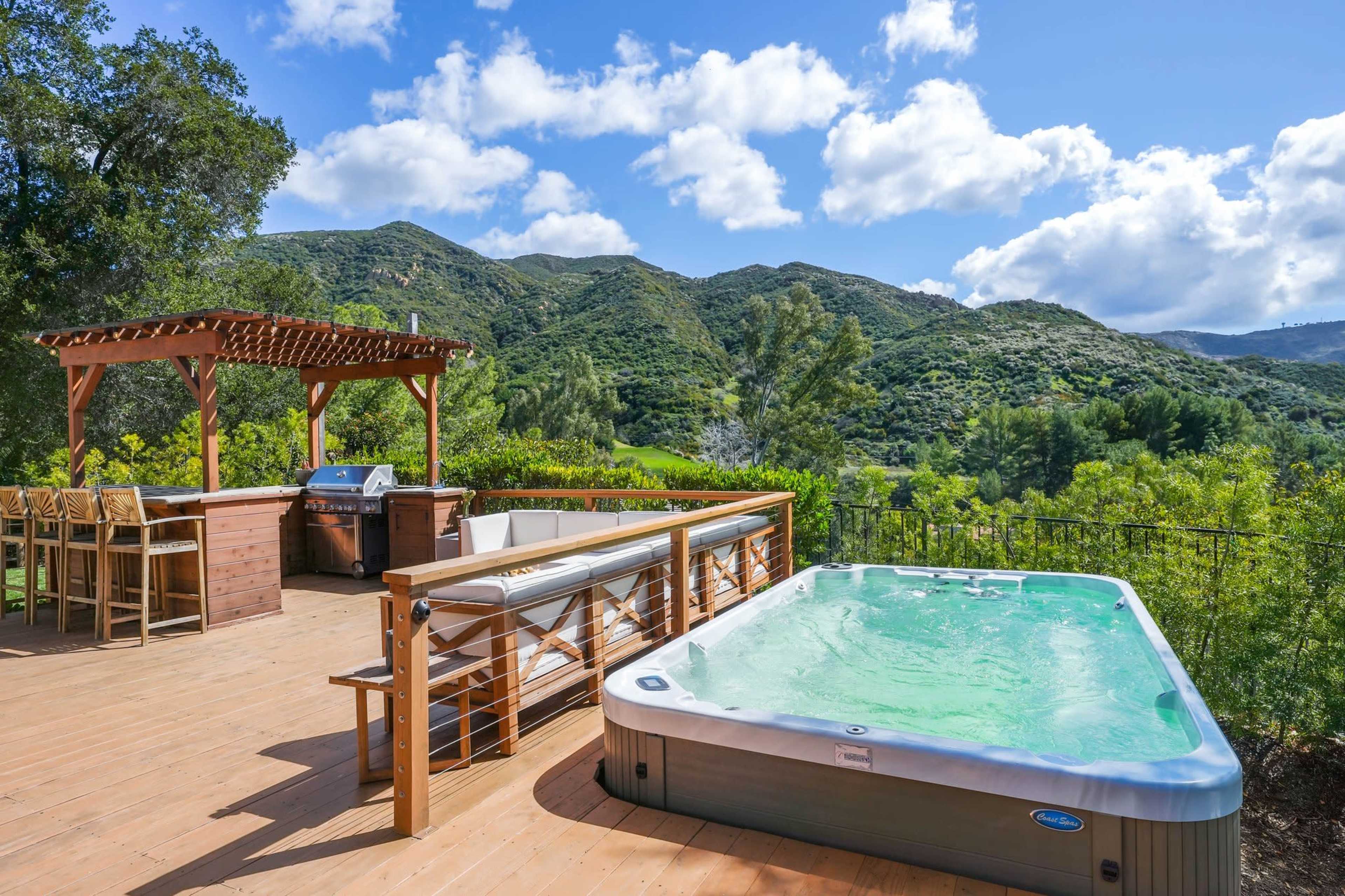 A wooden deck features a hot tub and a barbecue grill, surrounded by lush green hills and a clear blue sky.