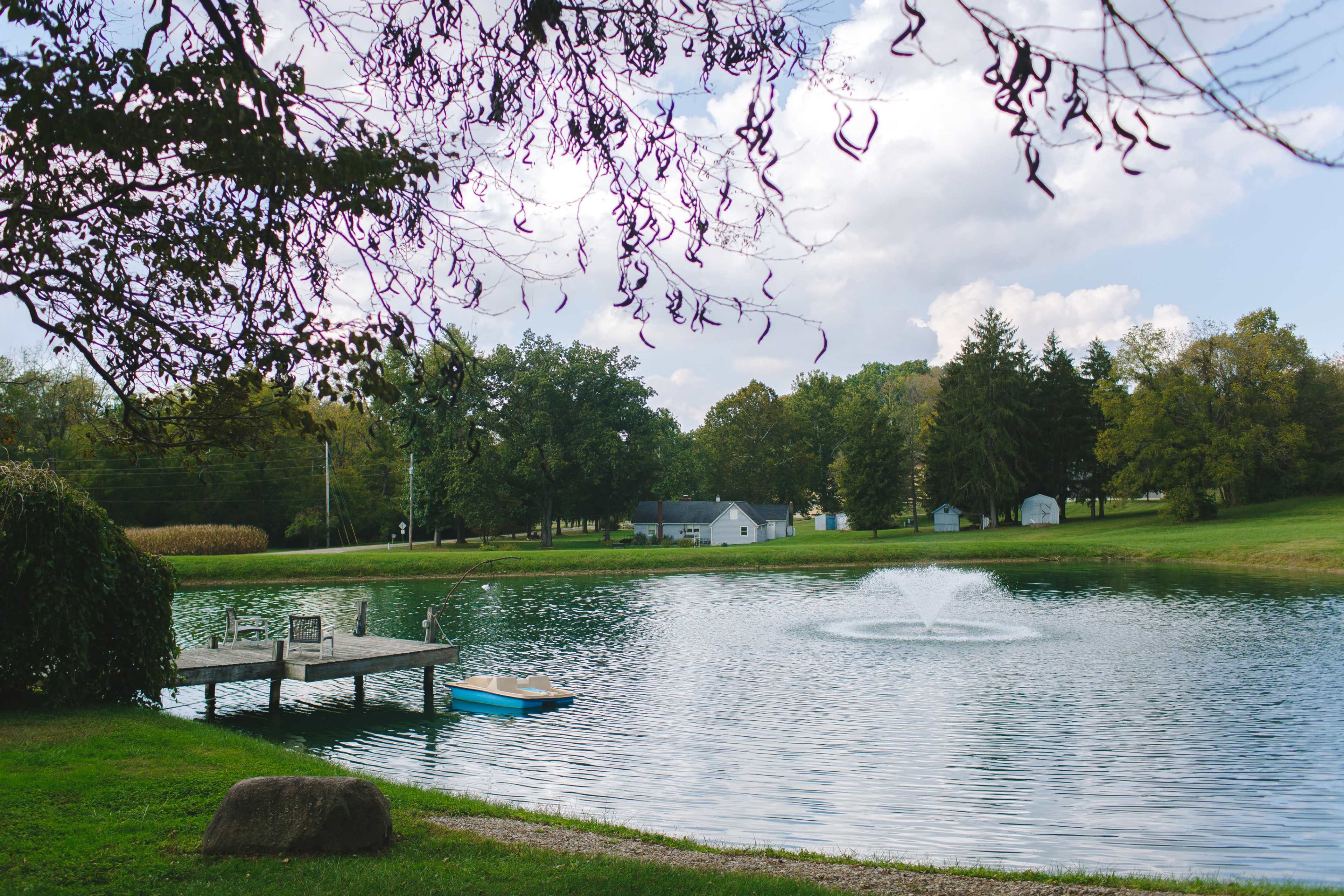 A small dock with chairs is situated beside a pond featuring a fountain, surrounded by trees and distant houses.