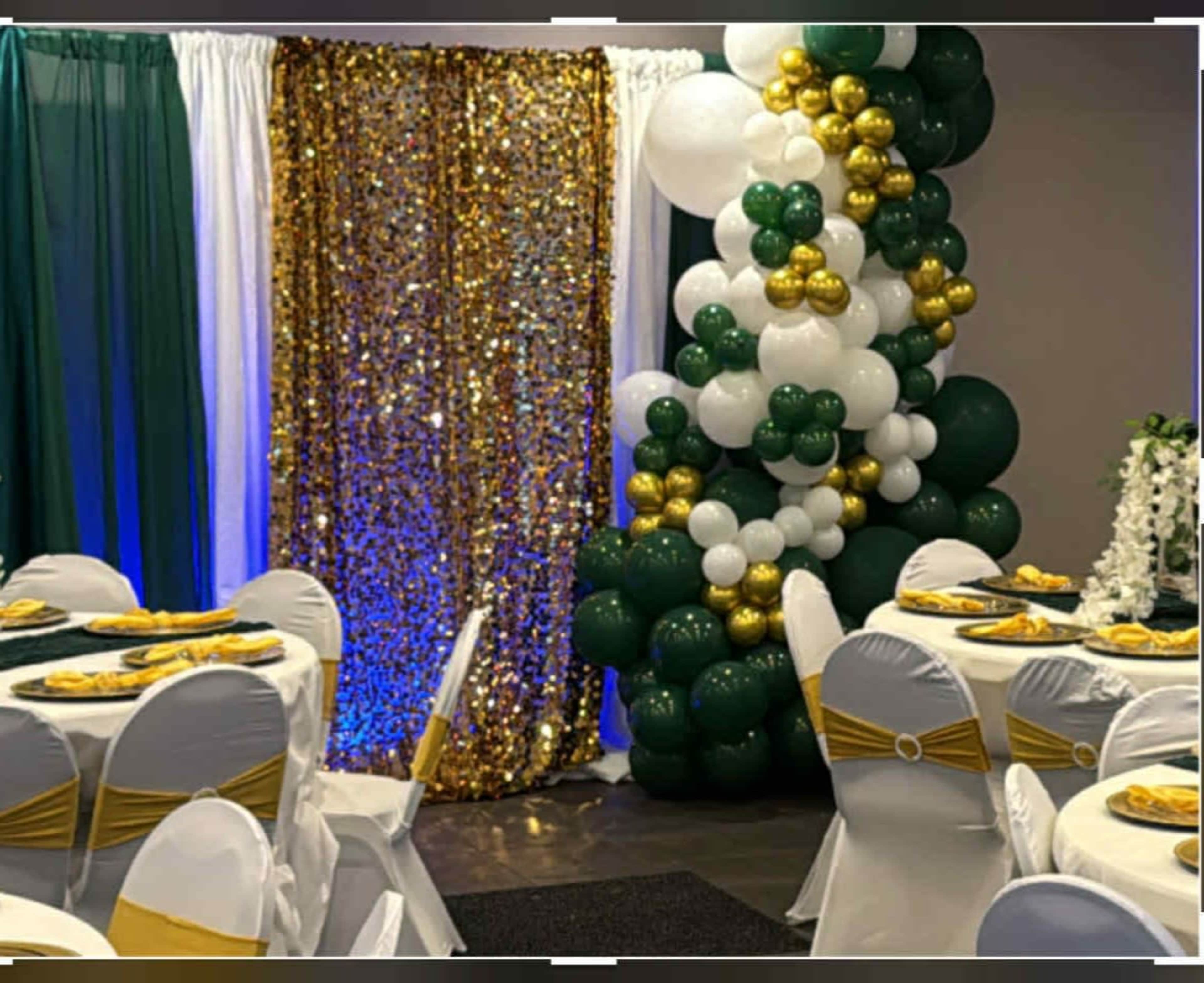The image shows a decorated event space featuring seated tables with gold and white table settings, a sequin backdrop with gold and green accents, and a large arrangement of balloons in similar colors.