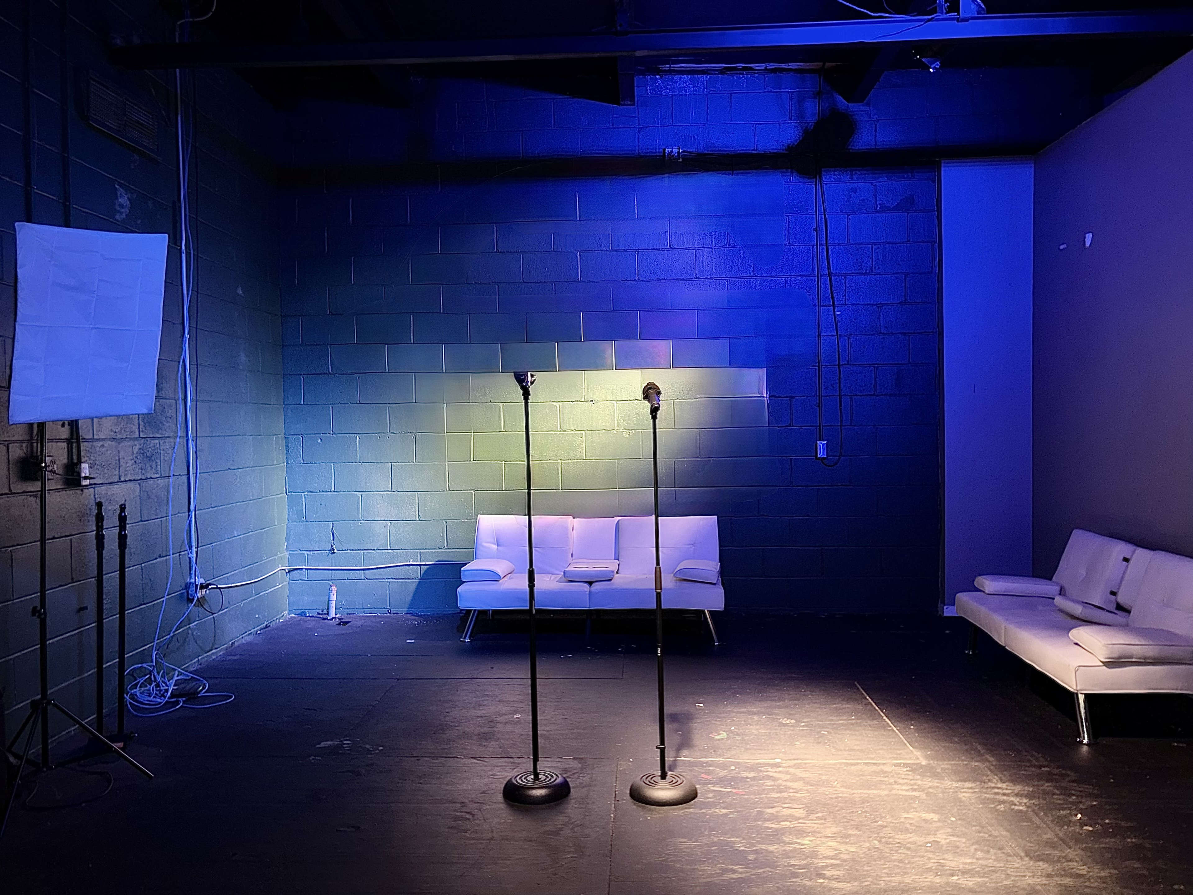 The image shows an empty stage with two microphones on stands and a white couch against a painted brick wall, illuminated by blue lighting.