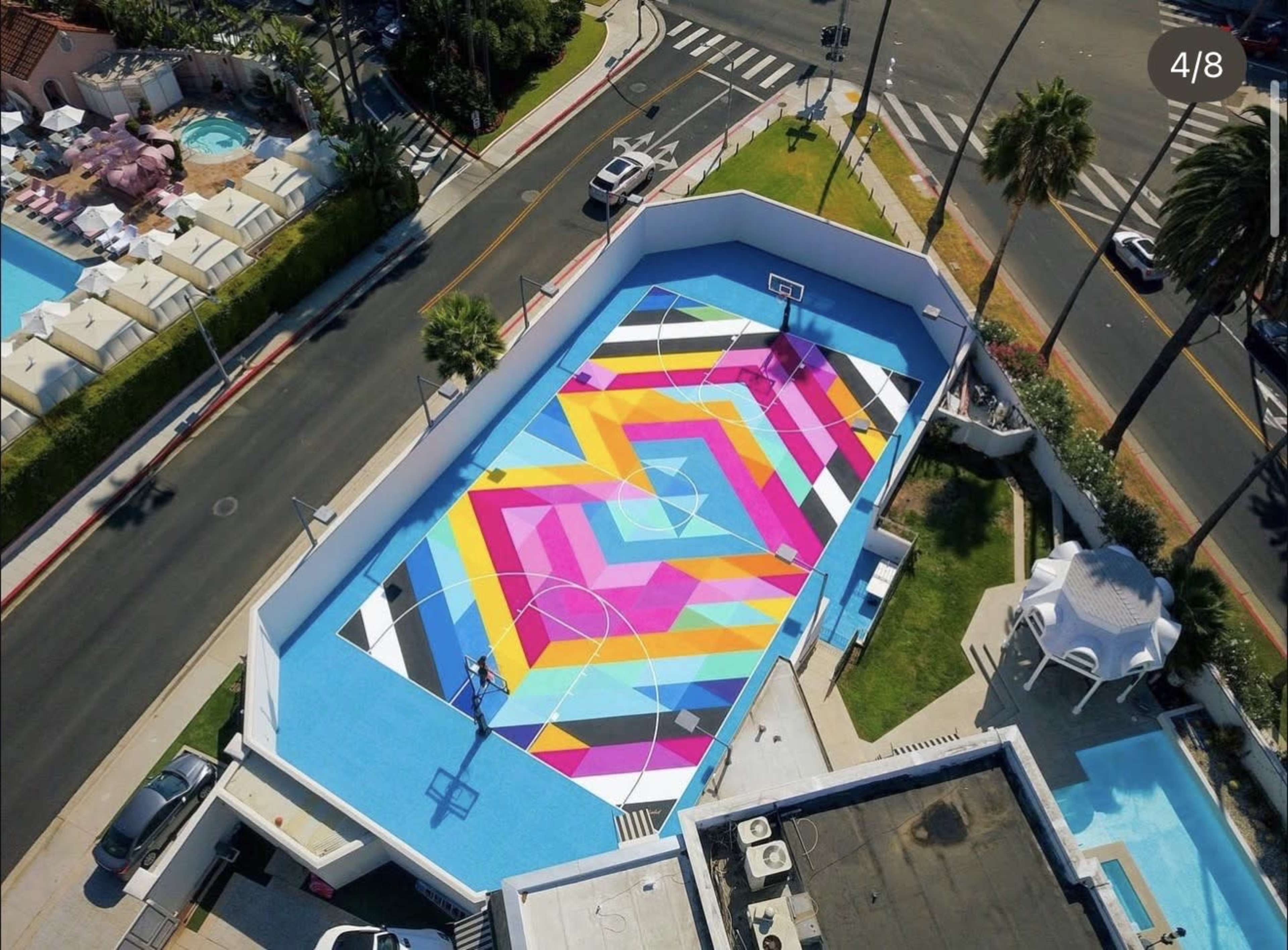 The image shows an aerial view of a colorful basketball court surrounded by palm trees and a swimming pool.