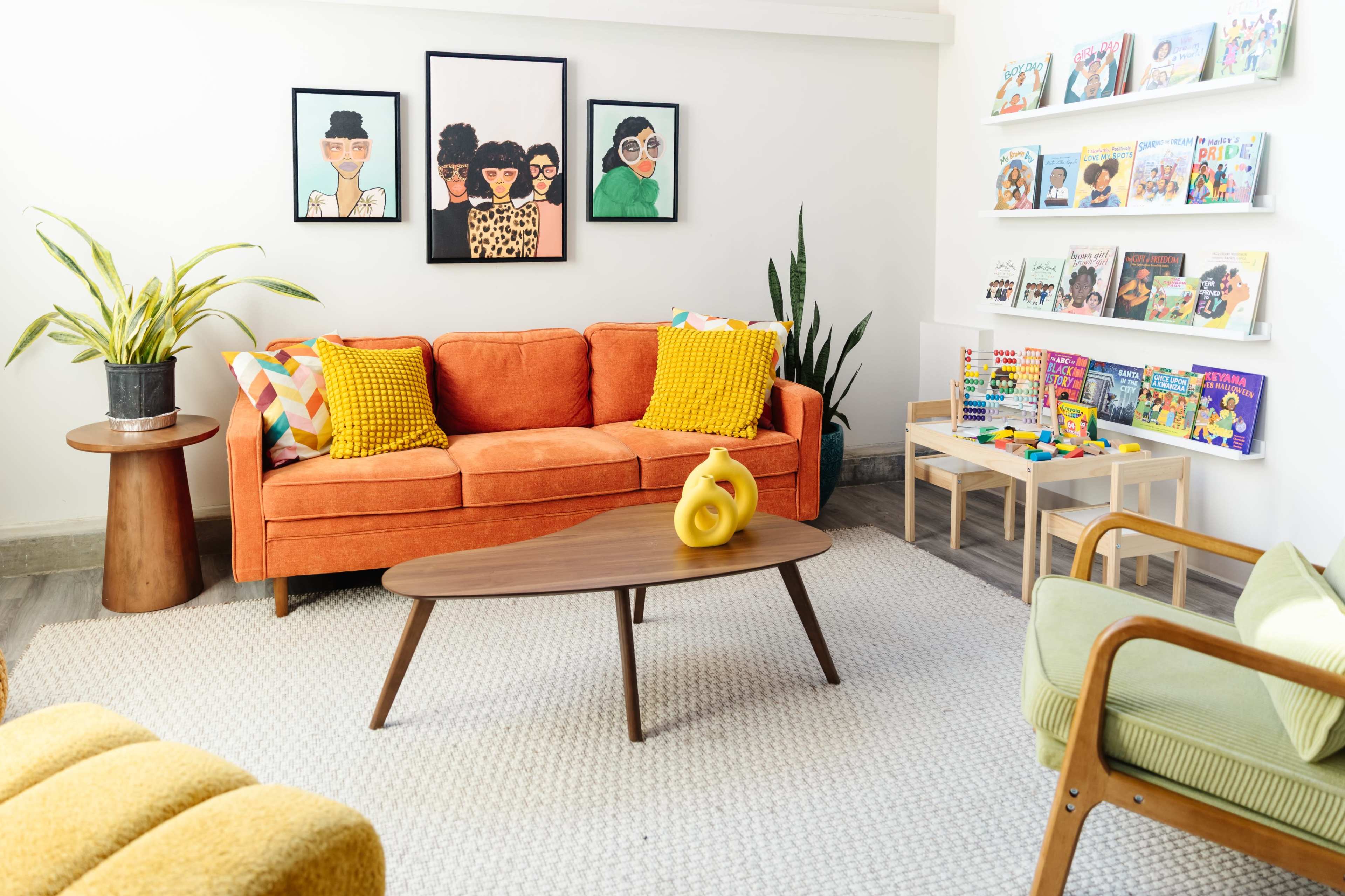 The image depicts a bright and inviting living room with an orange couch, a round wooden coffee table, colorful artwork on the walls, and books displayed on shelves.