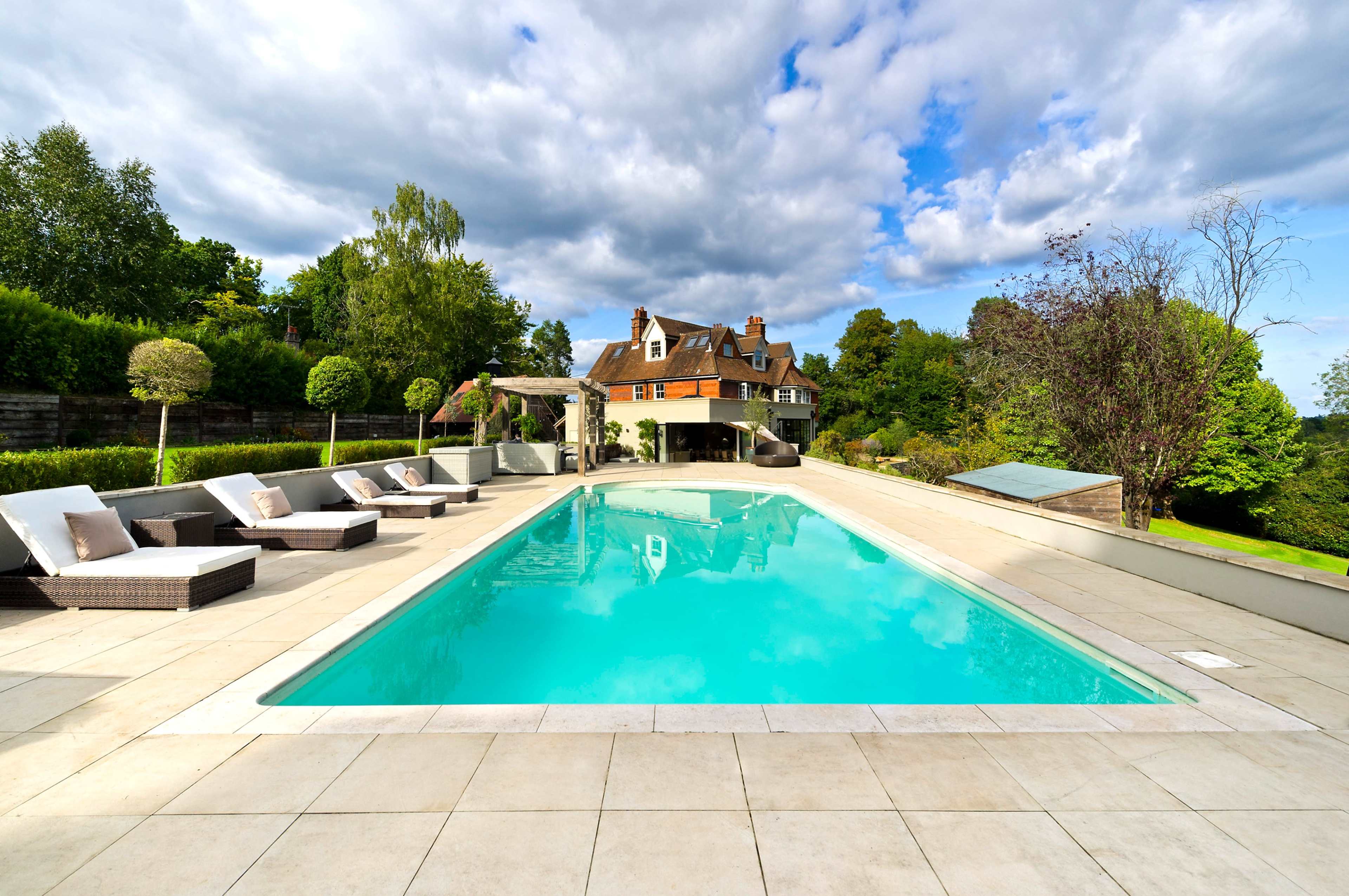 A rectangular swimming pool is surrounded by lounge chairs and overlooks a large house set against a backdrop of trees and cloudy skies.