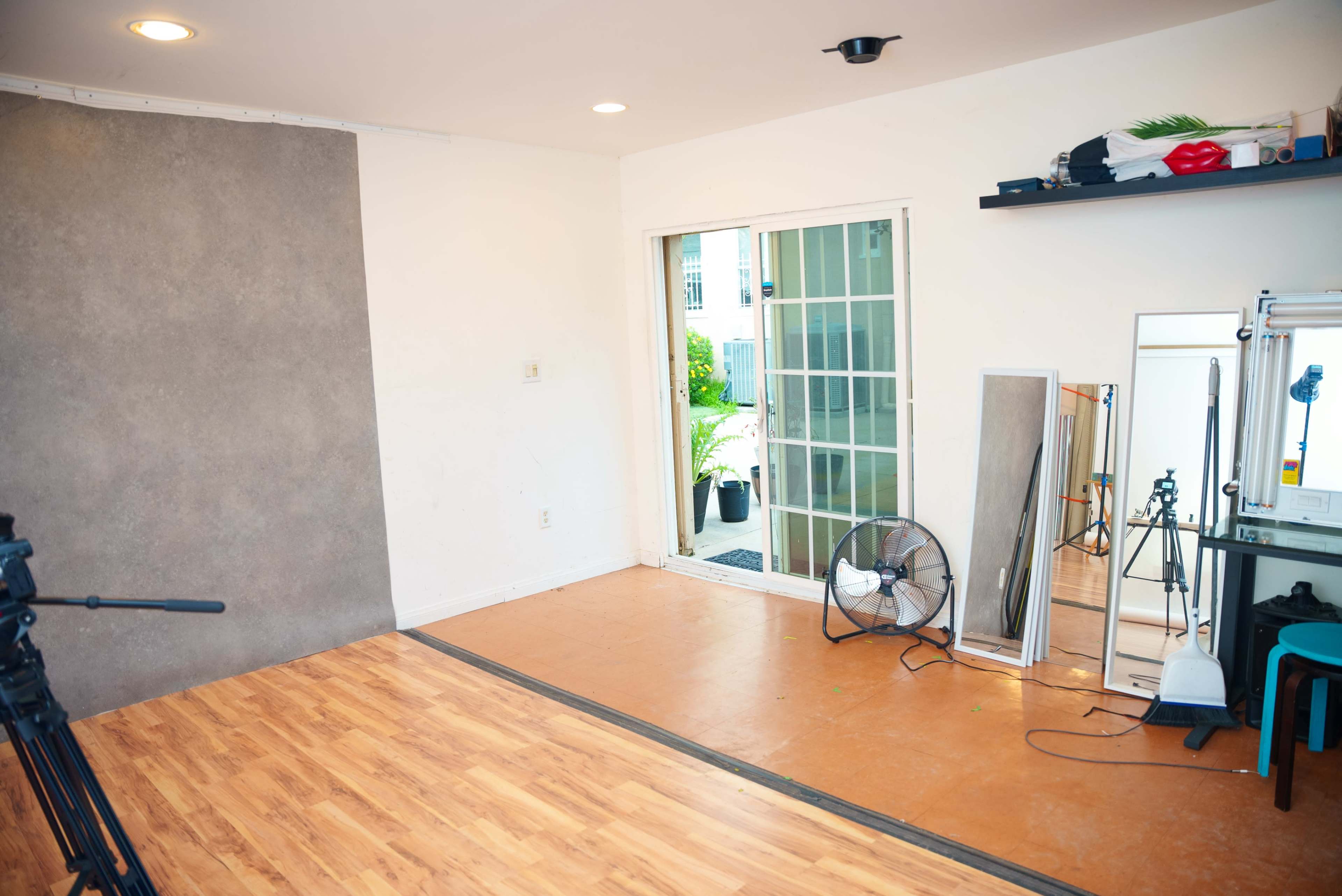 The image depicts an empty room with wooden flooring, a large window leading to an outdoor area, and various items including a fan, mirrors, and a shelf.
