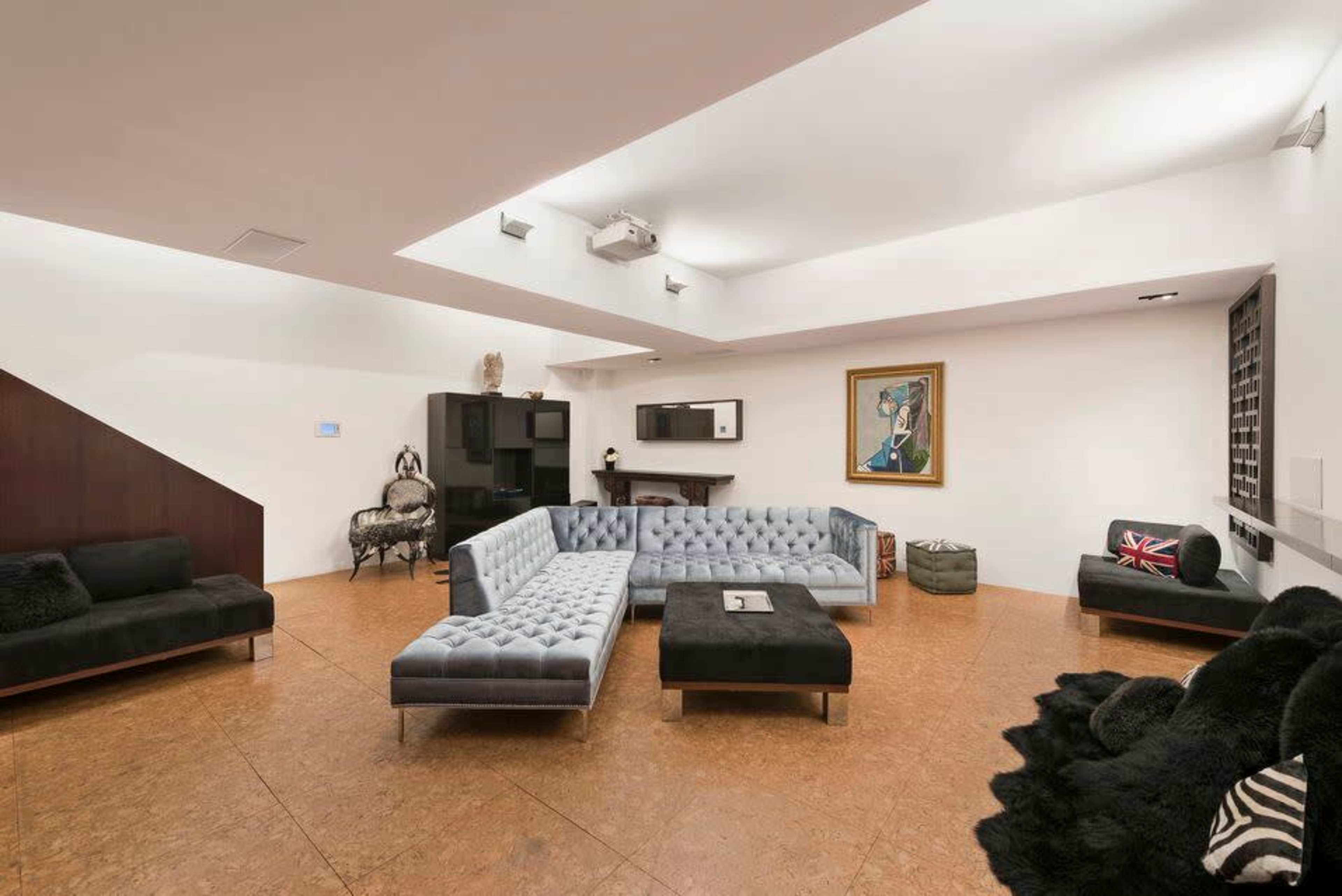 A spacious living room features a sectional sofa, a coffee table, and artwork on the walls, with a projector mounted on the ceiling.