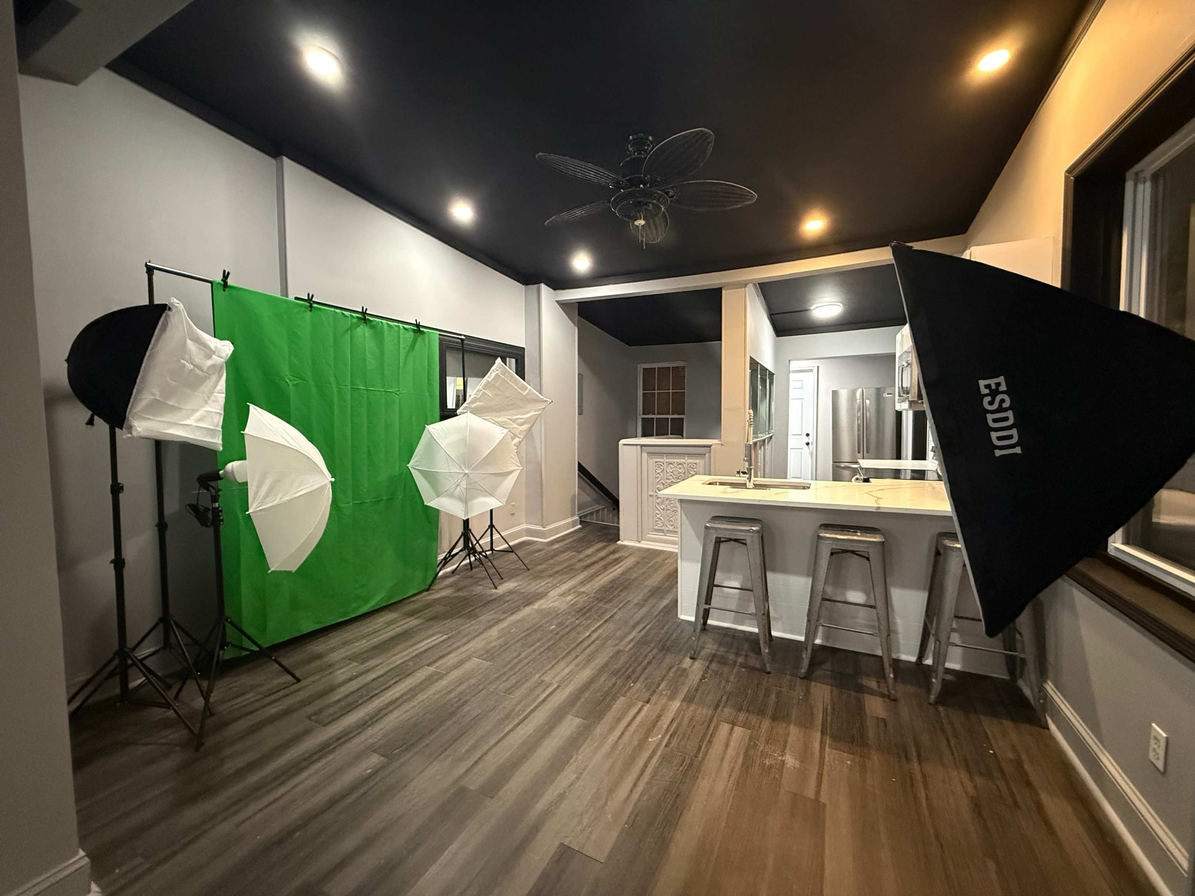The image shows a well-lit studio space featuring a green screen backdrop and several photography lights set up around a kitchen area with bar stools.