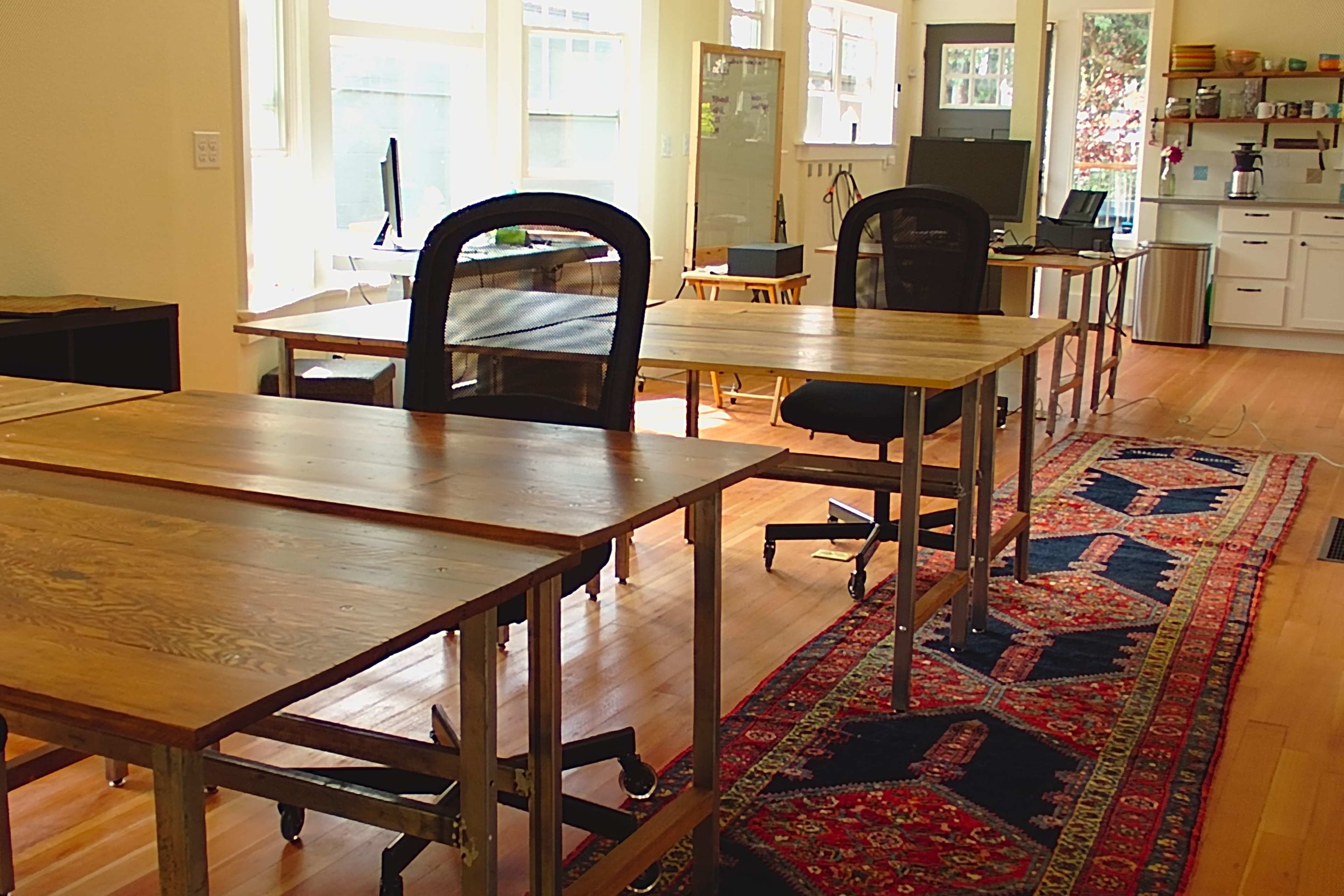 A spacious room with several wooden desks and chairs arranged on a patterned rug, adjacent to a kitchen area with white cabinets.