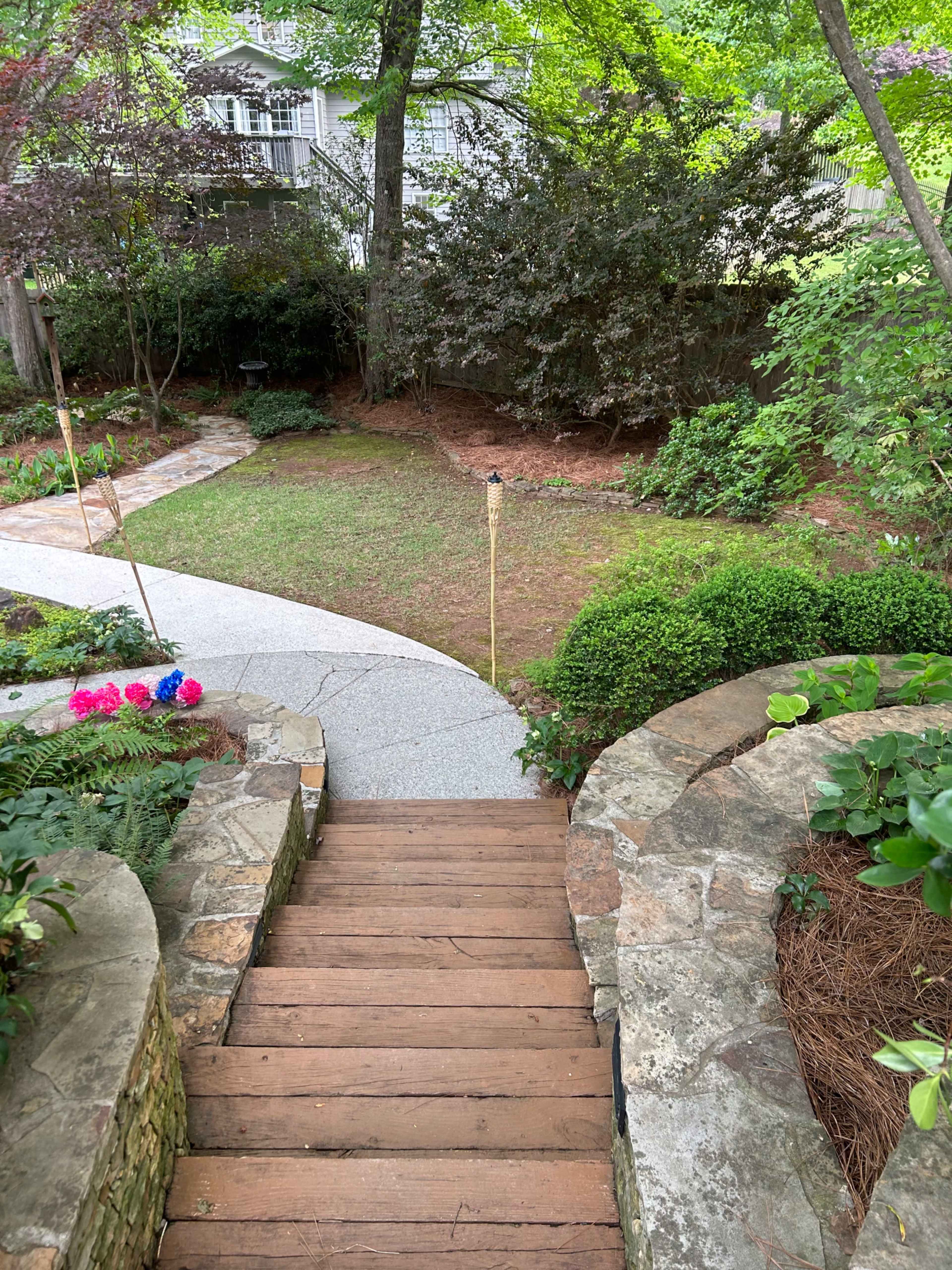 The image shows a wooden staircase leading down to a landscaped garden with a curved pathway, surrounded by greenery and decorative plants.